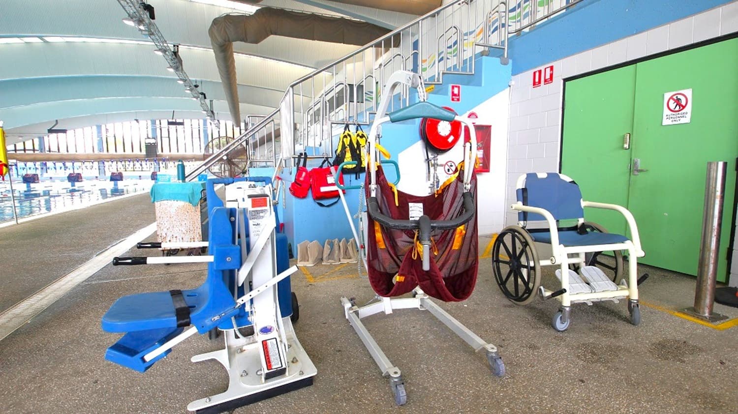 On the left, a blue pool hoist sits near the pool edge. In the middle, a patient lift with a maroon sling is positioned in front of a stairway with safety equipment and life vests hung on the wall. On the right, a blue and white aquatic wheelchair stands beside large green double doors. The floor is textured and the high ceiling has curved blue and white panels with lighting overhead.