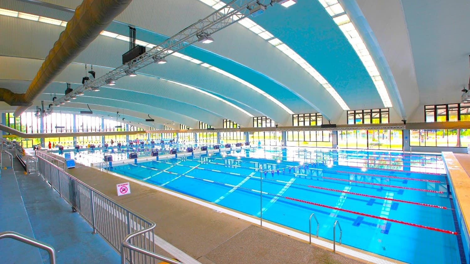 On the left, blue stadium seating stands behind stainless steel railings, adjacent to a wide beige poolside floor. The middle holds a large, clear swimming pool with racing lanes marked out. The right side features more poolside edge and tiled border. The background consists of rows of large windows, and the ceiling is arched with alternating blue and white panels, equipped with lighting and ventilation ducts.