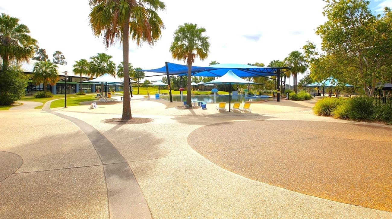 A spacious outdoor area with a smooth, curving pathway in the foreground. Several palm trees are scattered throughout the scene. In the middle, there are blue shade structures providing cover for tables and chairs. The background features more trees and a grassy area. The sky is partly cloudy, and the overall setting appears to be a park or recreational area.
