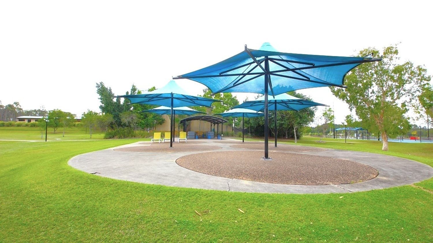 Large blue shade structures with black poles are positioned over a circular area with a textured surface. Surrounding the circle is a paved path, bordered by well-maintained grass. In the background, there are trees and a small building with a roof. To the right, a fence and more trees are visible. A few yellow chairs are placed under the shades.