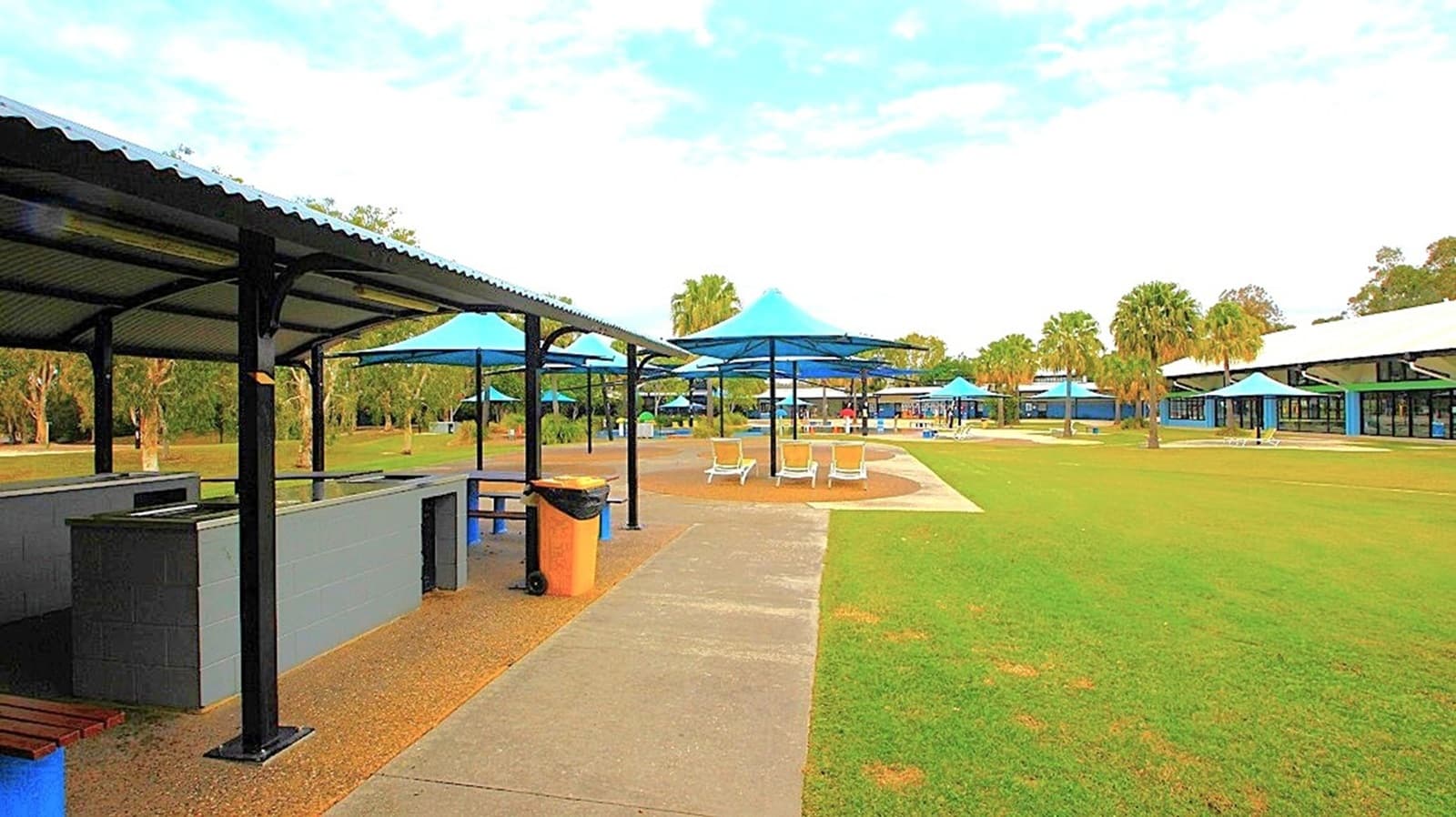 A park area with a paved walkway running through the centre. On the left, there are covered picnic areas with metal roofs and a bin nearby. To the right, a large grassy field is visible, with several blue canopies providing shade over seating areas. In the background, more canopies and a building with large windows can be seen. Trees are scattered throughout the area, adding greenery to the scene.