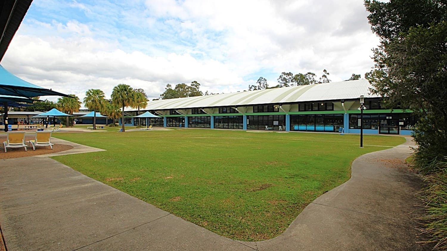 A spacious outdoor area with a large grassy lawn in the centre. On the left, there are several sun loungers and a pathway leading to a building with blue canopies. In the background, a long building with a white roof and blue pillars stretches across the scene. Palm trees are scattered around the area, and a few lamp posts are visible. The sky is overcast with some clouds.