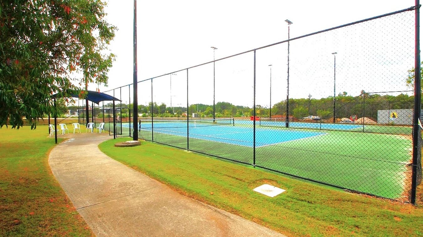 A tennis court with a blue playing surface is enclosed by a tall chain-link fence. On the left, a paved path runs alongside the court, leading to a shaded area with several white plastic chairs. Trees provide some greenery on the left side. In the background, there are more trees and some distant structures. The ground is covered with well-maintained grass.