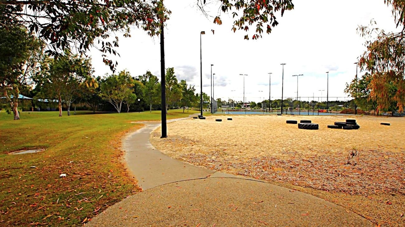 A park scene with a paved path curving through the foreground. On the left, there's a grassy area with scattered trees. To the right, a sandy area features several tyres arranged on the ground. In the background, tall floodlights and a fenced sports court are visible. The sky is overcast with clouds.