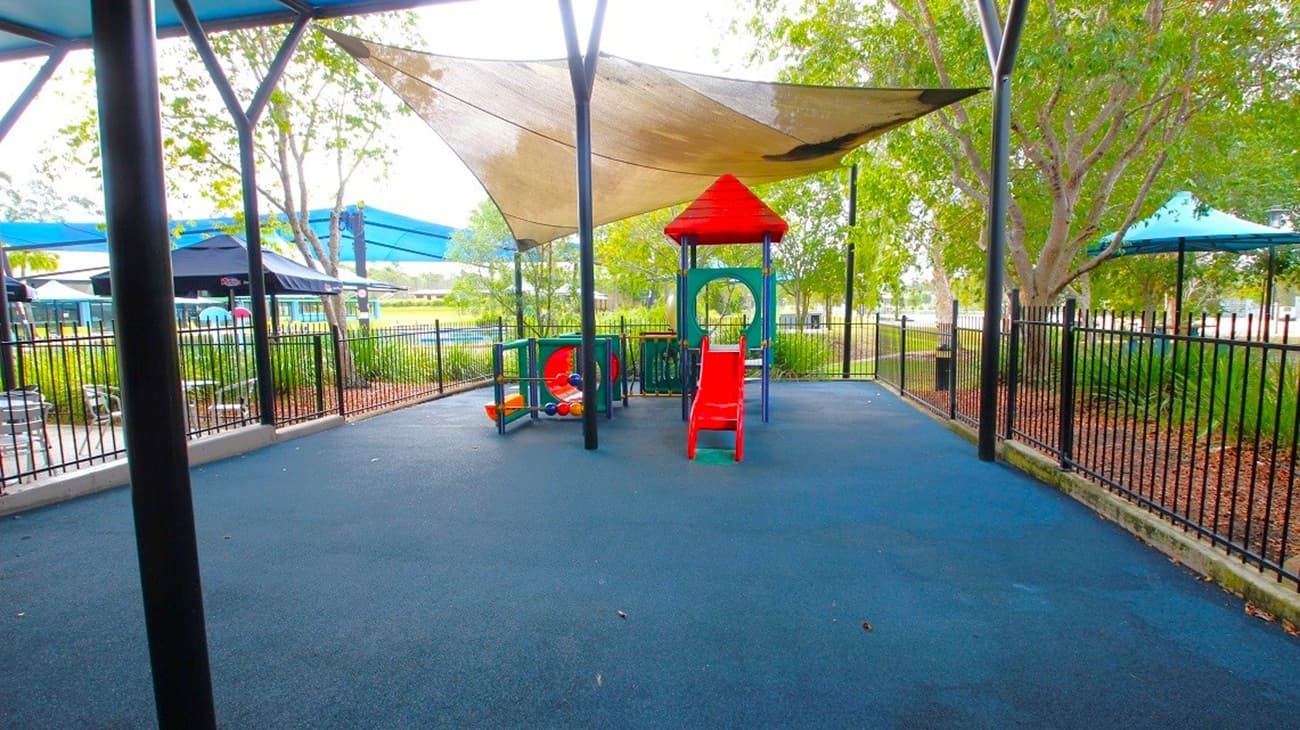 A small playground area with a blue rubber floor and a black metal fence surrounding it. In the centre, there is a play structure with a red slide and a small roof. A shade sail is overhead, supported by metal poles. Trees and shrubs are visible in the background, providing greenery. To the left, there are additional shaded areas with blue canopies.