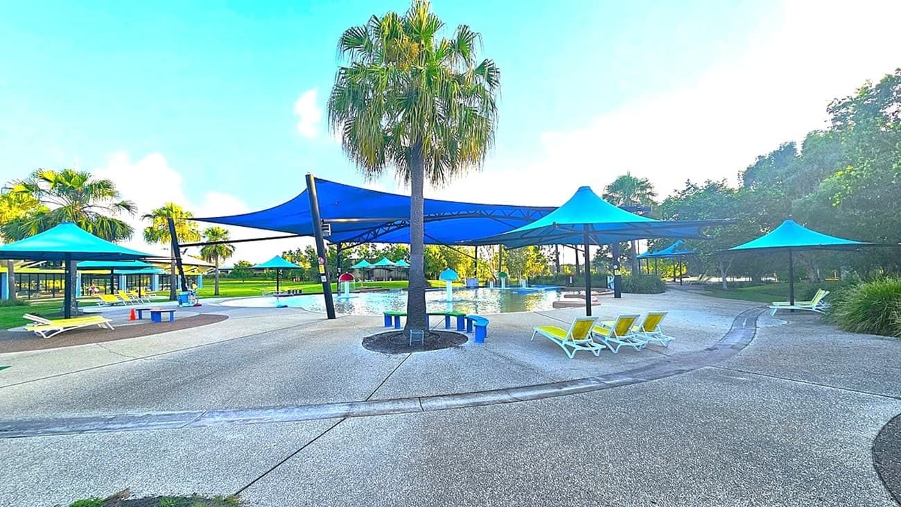 A spacious outdoor area with a central palm tree surrounded by a paved surface. Blue shade structures are spread across the area, providing cover over sections of the space. Yellow sun loungers are arranged on the right side, while colourful benches are on the left. In the background, there is a pool with more shade structures and trees lining the perimeter. The sky is clear, and the area is well-lit.