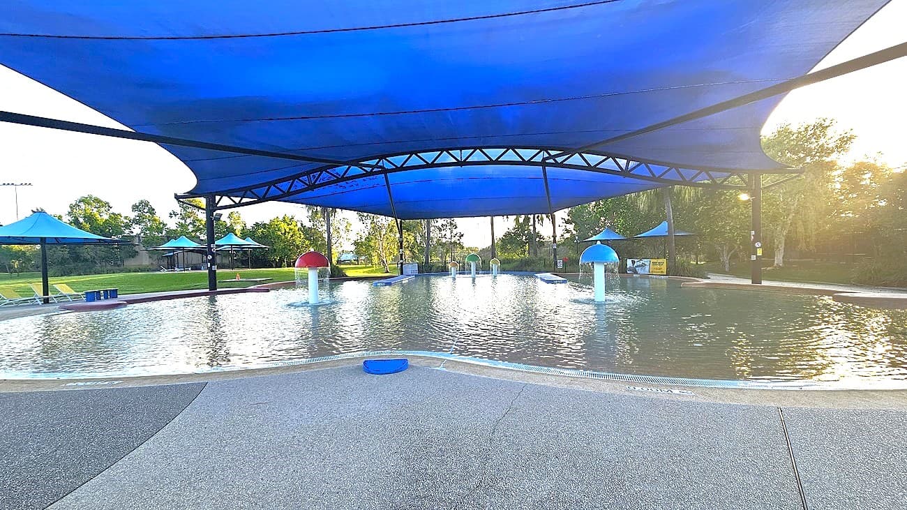 A shallow water play area with a textured concrete surface in the foreground. The pool features mushroom-shaped water fountains, one red and one blue, under a large blue shade canopy. Surrounding the pool are green lawns and trees. Blue umbrellas provide additional shade around the perimeter. The area is well-lit with sunlight filtering through the trees in the background.