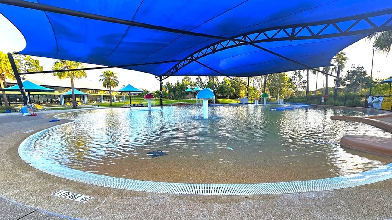 A shallow water play area with a textured concrete edge and a blue shade structure overhead. In the centre, there are several water features shaped like mushrooms, with water cascading from their tops. Surrounding the pool are grassy areas and palm trees. In the background, there are additional shaded areas with blue canopies and a building with a yellow roof. The pool's depth is marked as 0.00m on the edge.