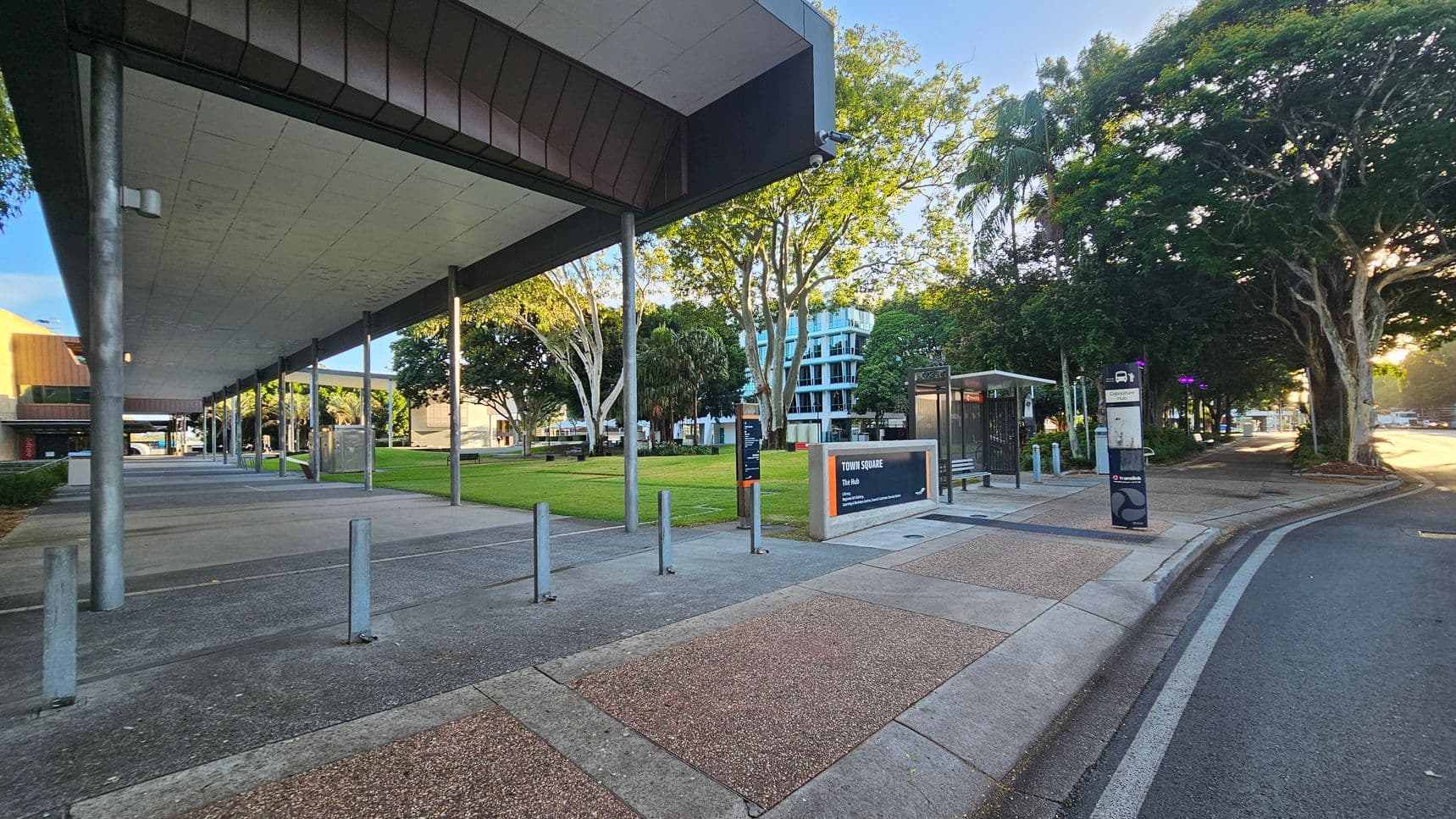 A modern urban area with a wide walkway on the left, covered by a large overhanging roof supported by metal poles. The ground is a mix of concrete and textured paving. In the centre, there's a grassy area with trees and a sign reading "Town Square." On the right, a bus stop with a shelter and a signpost is visible. The background features a multi-storey building partially obscured by trees. The scene is well-lit, suggesting early morning or late afternoon.