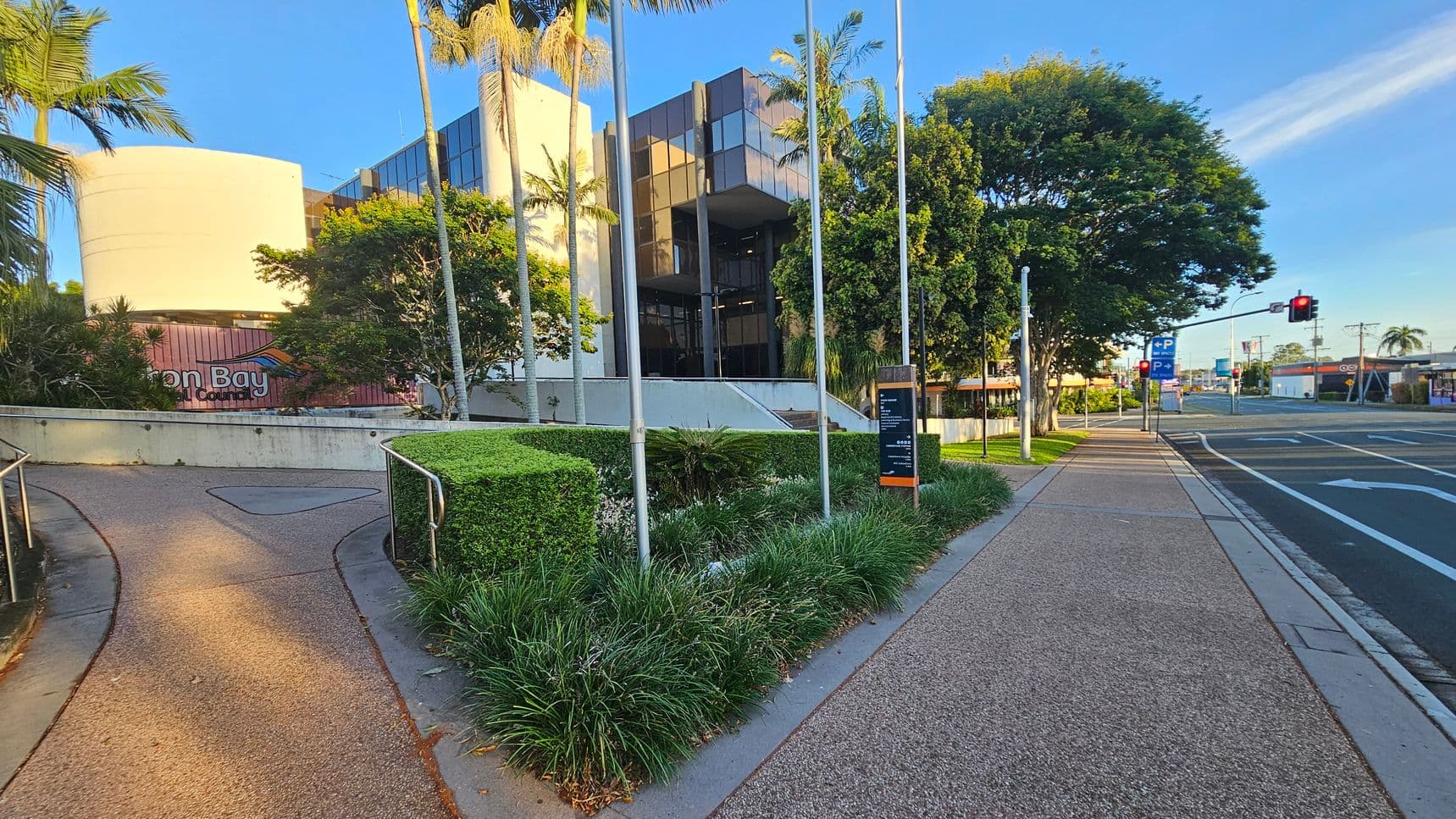 A modern building with reflective glass panels and a cylindrical white structure is in the background, surrounded by tall palm trees and lush greenery. In the foreground, a paved path curves to the left, bordered by manicured hedges and grass. On the right, a straight pavement runs alongside a road with visible traffic lights and parking signs. The scene is well-lit, suggesting daylight.
