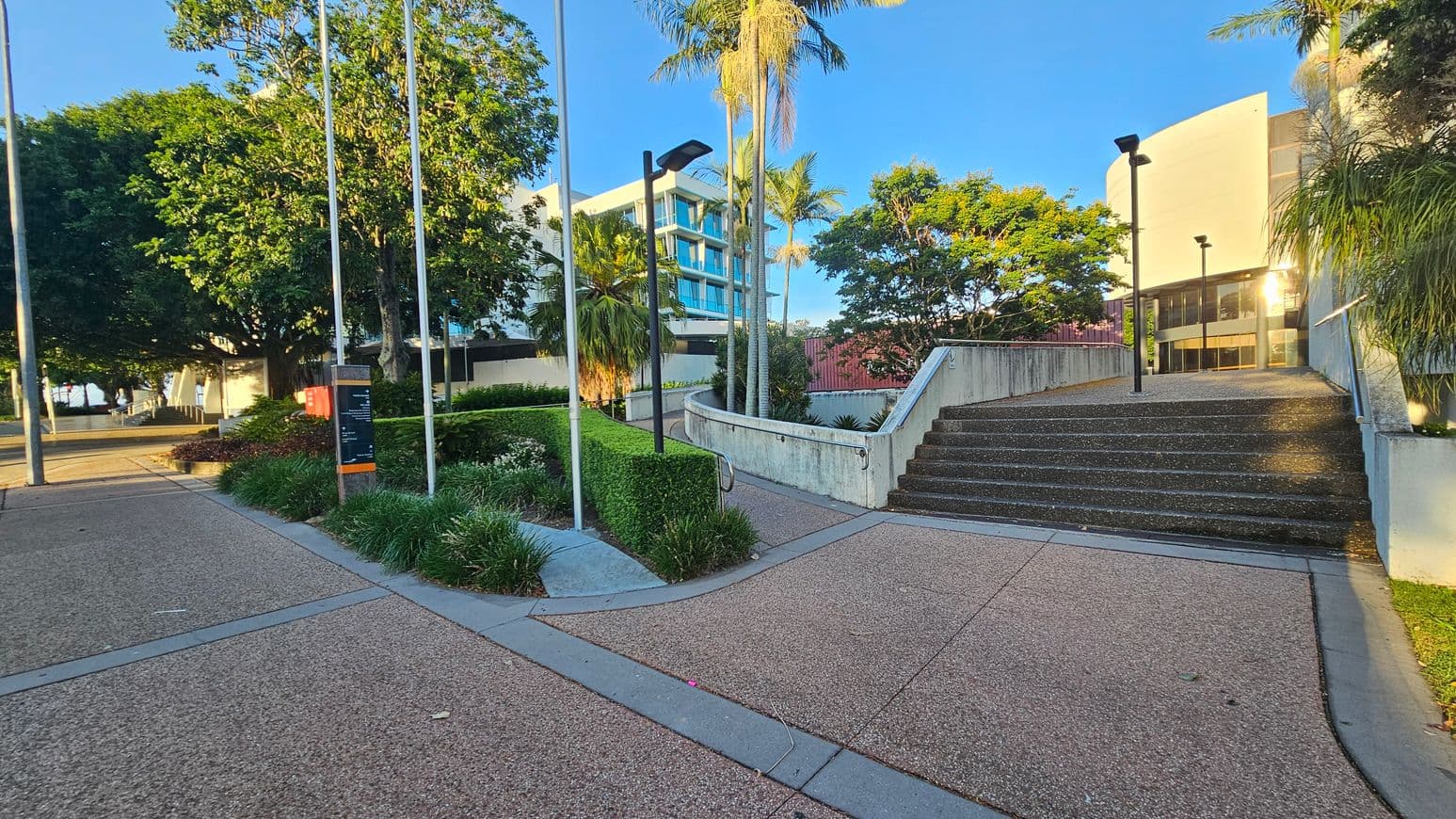 A paved area with a path leading to a set of stairs on the right. The stairs are bordered by a low concrete wall with metal railings. On the left, there are flagpoles and a sign near neatly trimmed hedges and various plants. Tall palm trees and a large leafy tree provide greenery. In the background, a modern building with blue-tinted windows is visible. The area is well-lit with street lamps, and the ground is covered with small pebbles.
