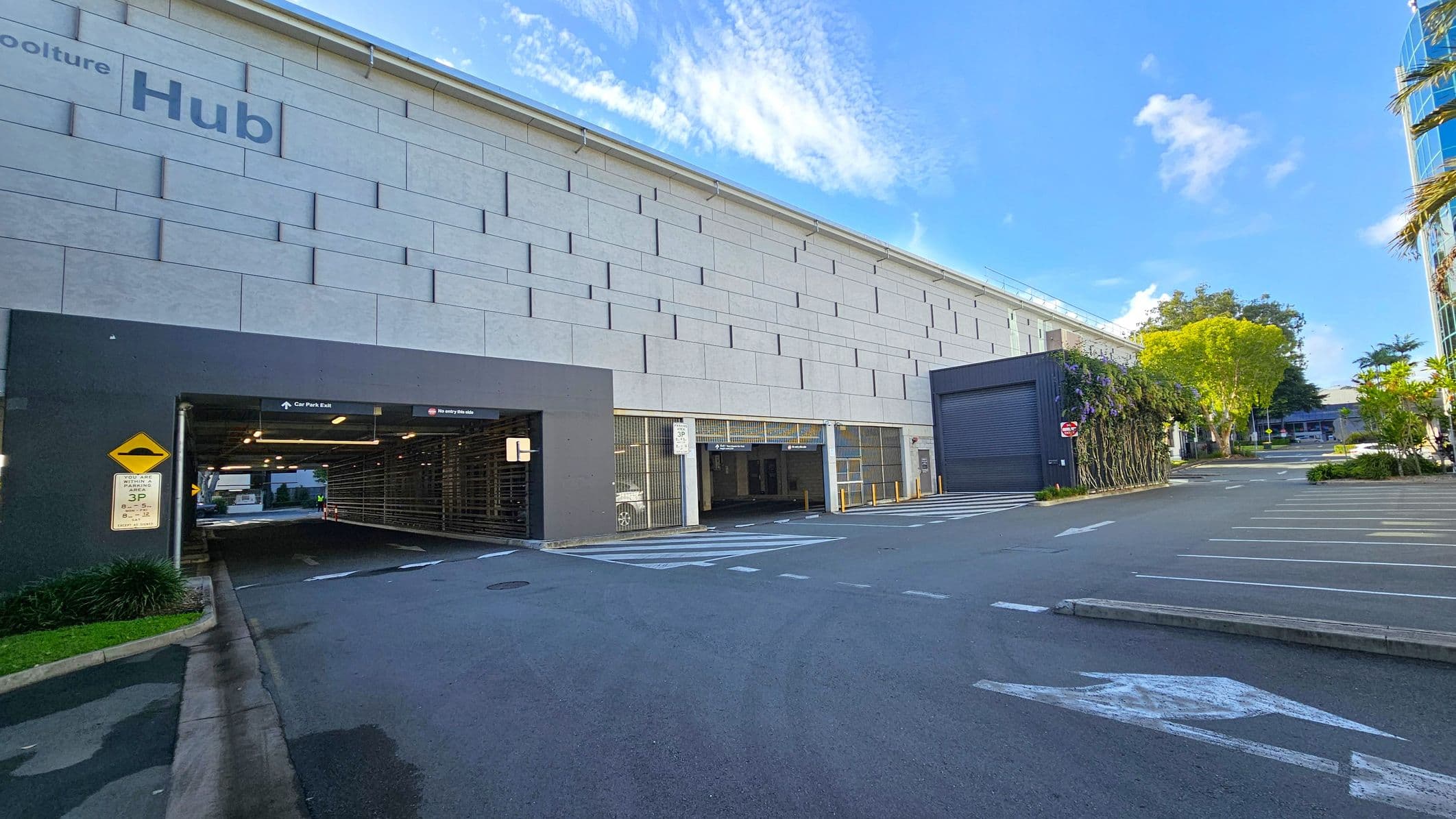 A large building with a grey, textured facade on the left, labelled "Hub". Below, there is an entrance to a car park with signs indicating directions. The road in front has marked parking spaces and directional arrows. On the right, a section of the building is covered with greenery, and a tree is visible further along the road. The background includes more trees and a glimpse of another building. The ground is paved, and the sky is clear.
