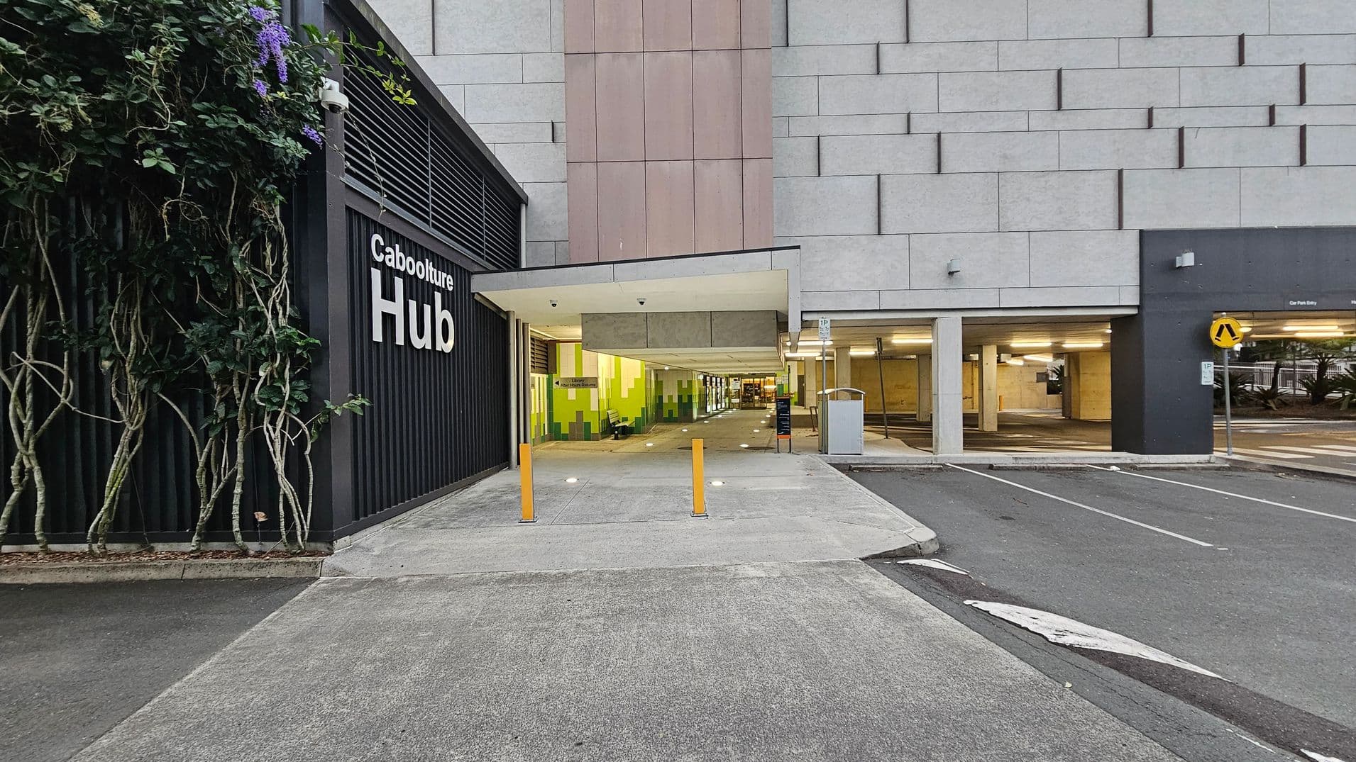 On the left, a black wall with vertical slats and climbing plants features the words "Caboolture Hub". In the centre, a covered walkway with a slanted roof leads into a building with green and yellow walls. Three yellow bollards are positioned at the entrance. On the right, a car park entry is visible with a pedestrian crossing sign. The ground is paved with concrete, and the building's exterior is clad in grey panels.