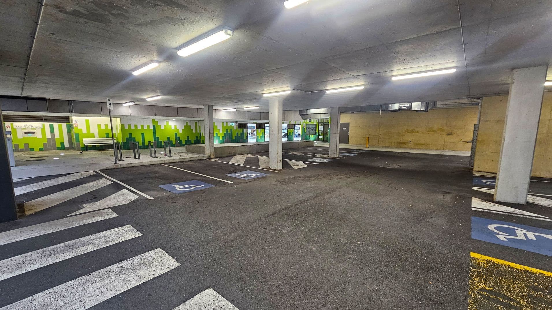 A covered car park with several parking spaces marked for disabled access, indicated by blue paint and wheelchair symbols. The floor is asphalt with white zebra crossing lines. The ceiling is concrete with fluorescent lights. The background features a wall with a green and white geometric pattern, and a bench is visible. Several concrete pillars support the structure, and there are signs and doors along the back wall.