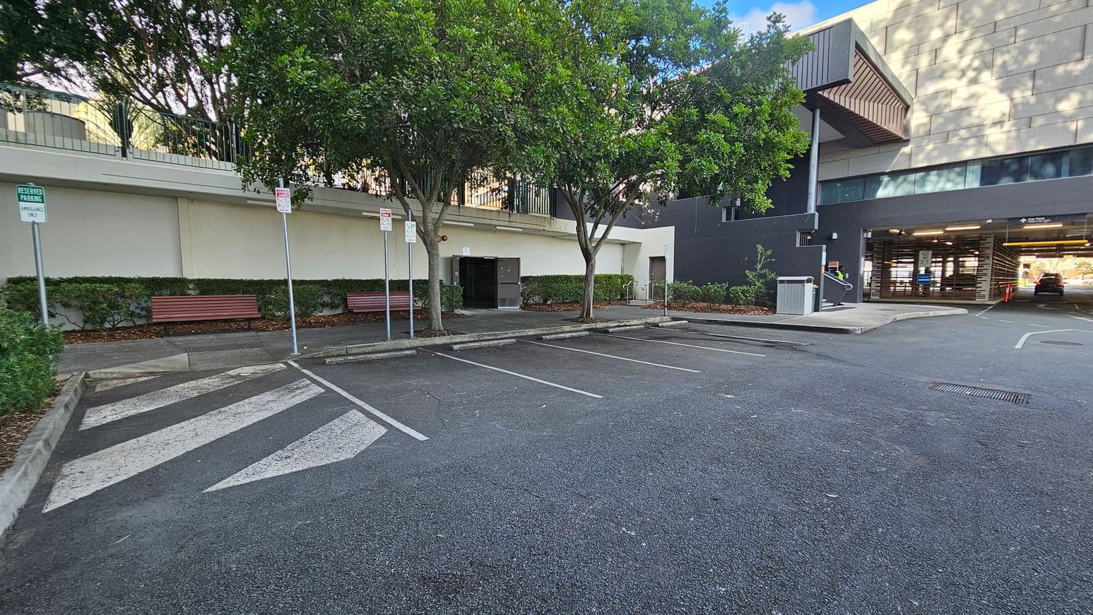 A parking area with reserved spaces marked by signs on the left, including one for ambulances. Two wooden benches are positioned along the wall, with trees providing shade. The ground is asphalt with painted lines and a pedestrian crossing. On the right, a building with a purple wall and an entrance to a covered parking area is visible. Bushes line the wall, and a small ramp leads to a door.