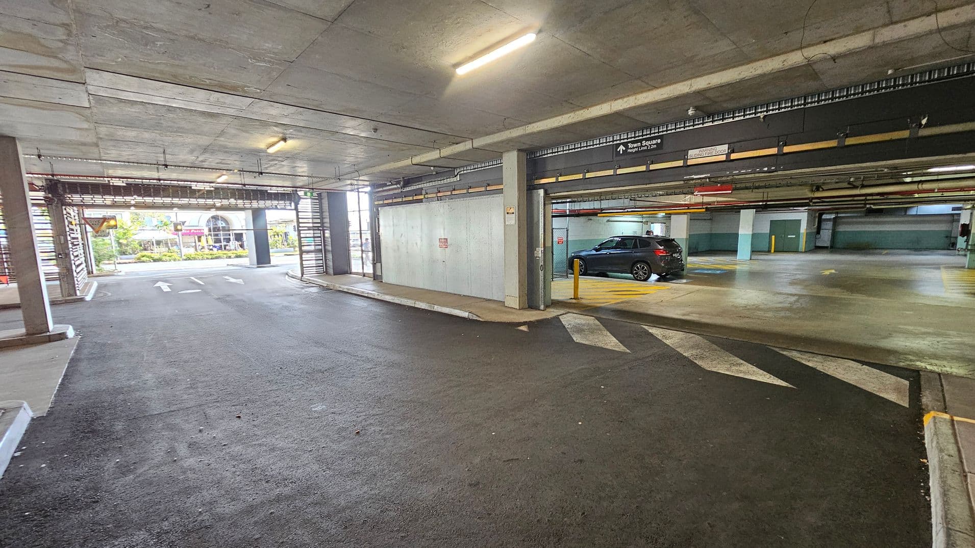 A multi-storey car park with a concrete ceiling and floor. On the right, a black car is parked in a designated space with yellow markings. The left side shows an exit with arrows on the ground leading outside, where a building and greenery are visible. Overhead, fluorescent lights illuminate the area. A sign above the car park entrance indicates directions to "Town Square" and height limits.