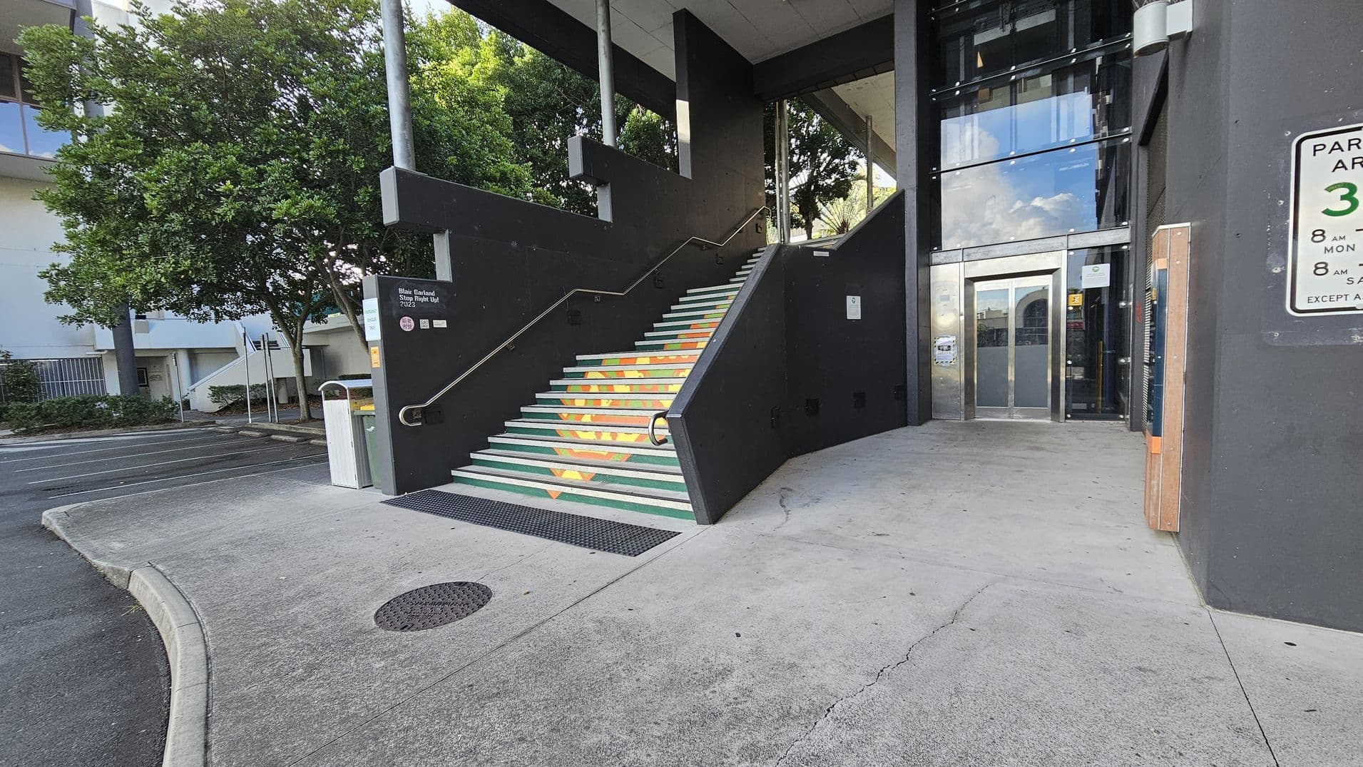 Concrete stairs with colourful geometric patterns lead up to a building entrance on the right. The entrance has glass doors and is surrounded by dark walls. A parking sign is visible on the right wall. On the left, there is a tree and a small parking area. The ground is paved with concrete, and a metal pole stands near the stairs. The ceiling above the entrance is flat and dark.
