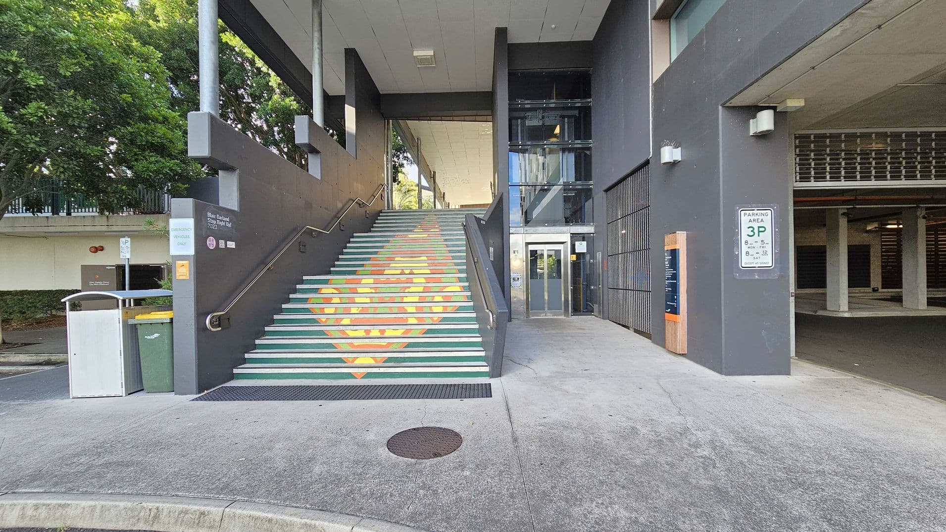 A concrete staircase with colourful geometric patterns leads up to a covered area. To the right, there is a glass lift with a metal frame. A parking sign is mounted on the wall near the entrance to a parking area. On the left, a green bin and a yellow-lidded bin are placed next to the stairs. Trees and a fence are visible in the background. The ceiling is flat and grey, and the ground is paved with concrete.
