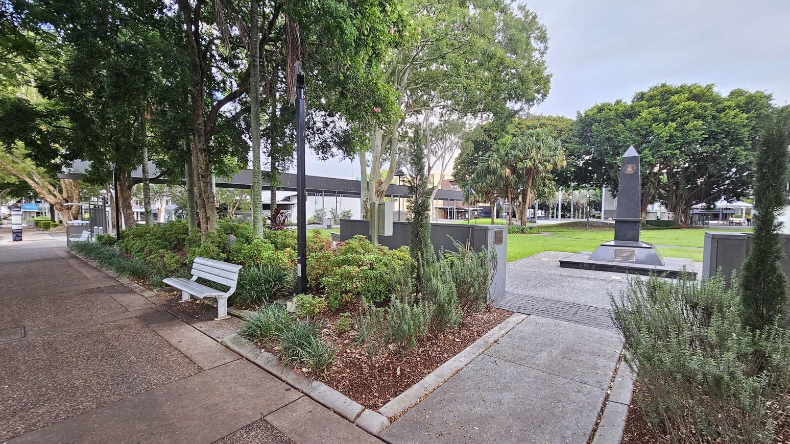 A park scene with a paved path on the left, featuring a white bench. To the right, there's a garden area with various shrubs and trees. In the centre, a black obelisk stands on a platform surrounded by grass. In the background, more trees and a few buildings are visible. The ground is a mix of concrete and grass, and the area is well-maintained.