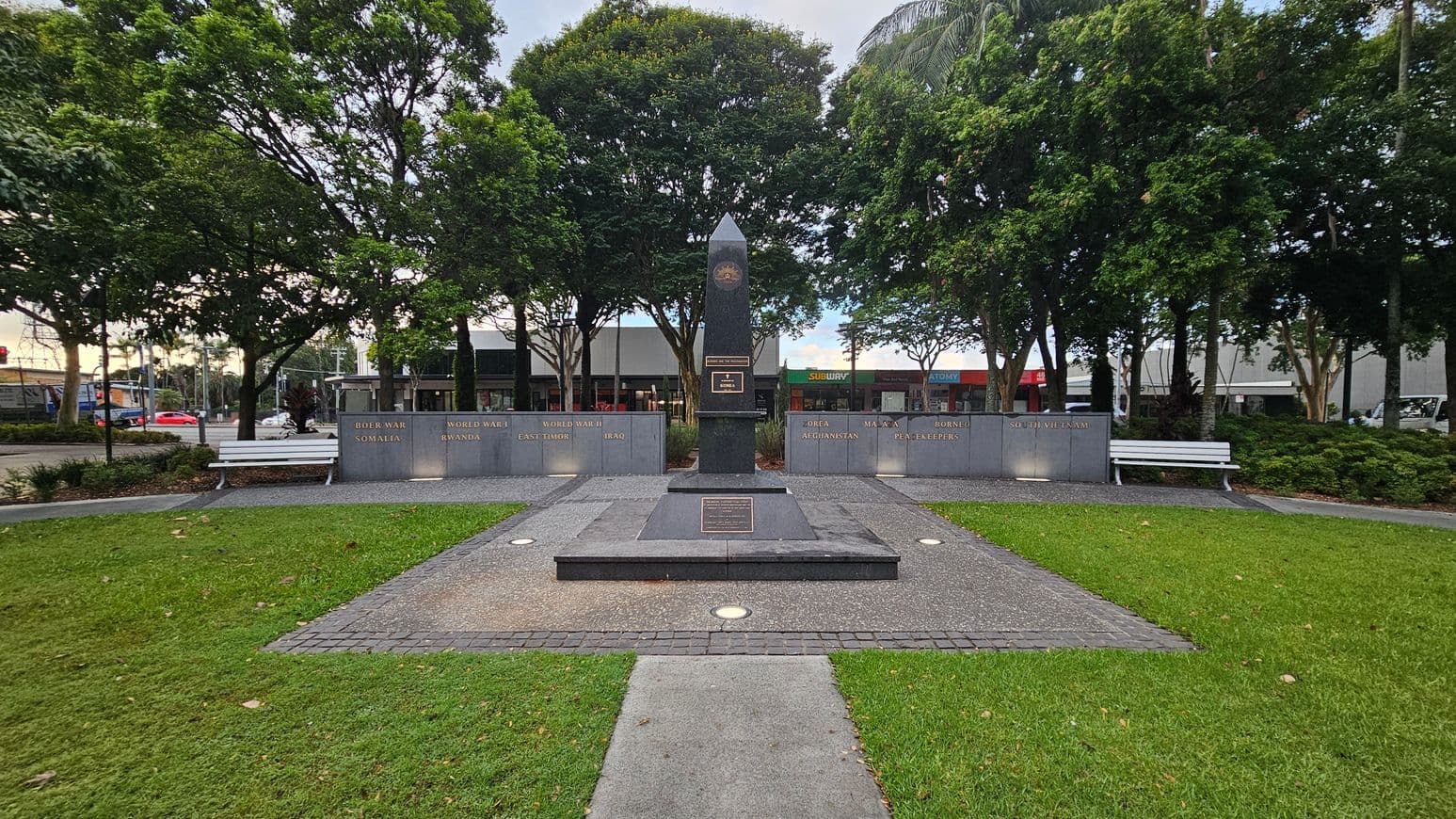 A war memorial with a central obelisk surrounded by plaques listing various conflicts, including the Boer War, World Wars, and others. Two white benches are positioned on either side. The area is paved with a path leading to the monument, bordered by grass. Trees form a backdrop, and a street with shops, including a Subway, is visible in the background.
