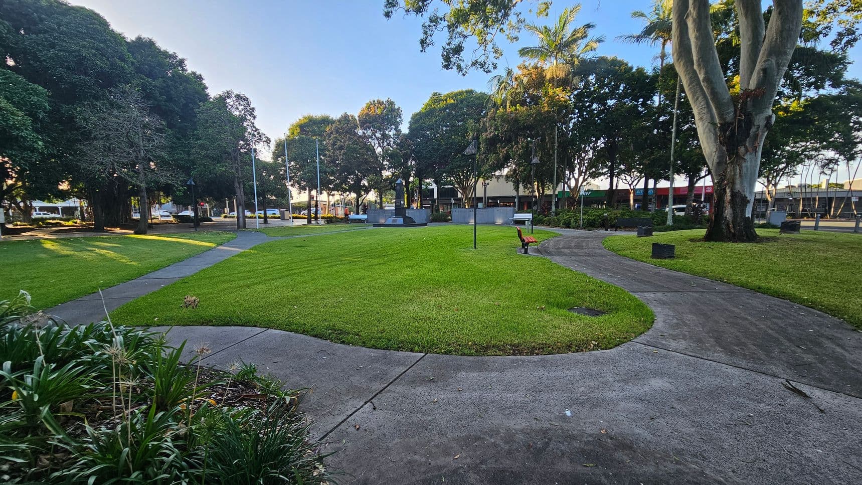 A park with a curved pathway leading through a grassy area. On the right, a large tree with a thick trunk stands near a bench. In the background, several flagpoles and a monument are visible, surrounded by more trees. To the left, there are additional trees casting shadows on the grass. Buildings and parked cars can be seen in the distance. The foreground features some low plants and a well-maintained lawn.