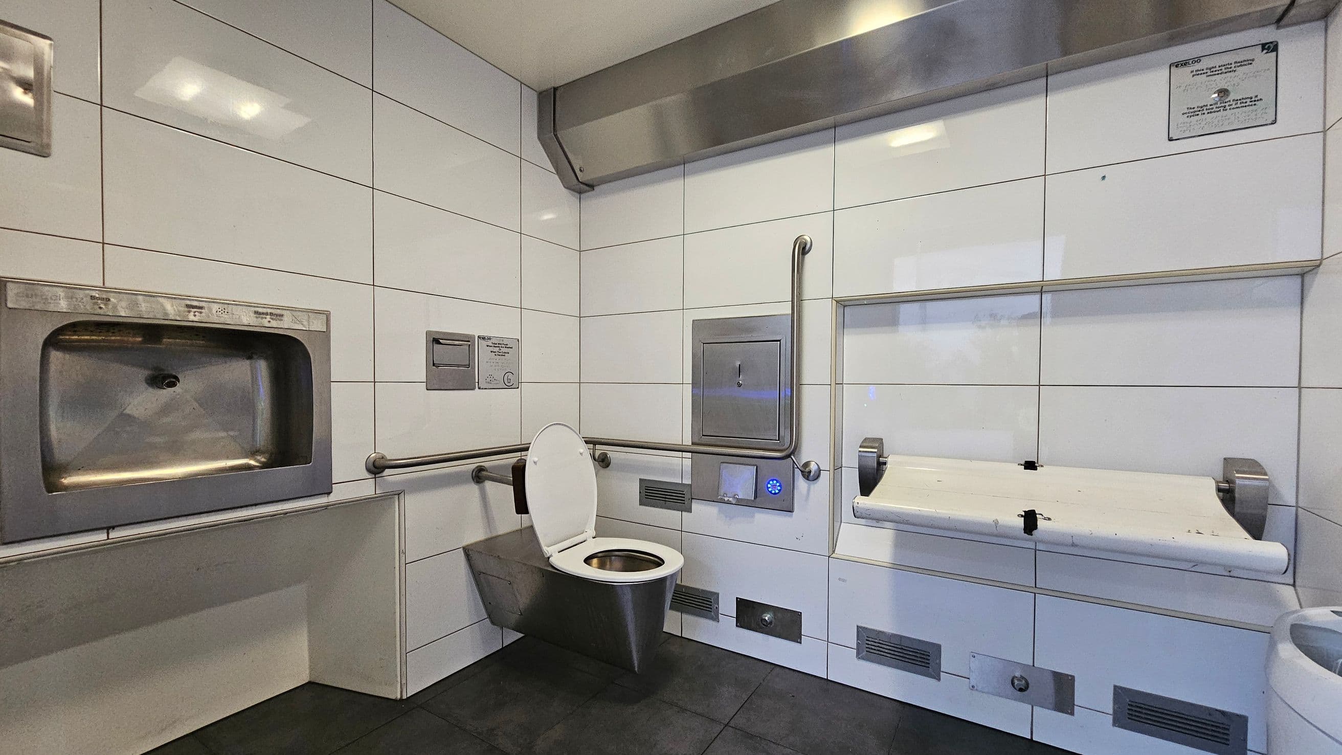 A public restroom with white tiled walls and a dark tiled floor. On the left, there is a stainless steel hand dryer. In the centre, a wall-mounted toilet with a white seat and grab bars on either side. Above the toilet, a metal panel and a small sign are visible. On the right, a foldable baby changing table is attached to the wall. A ventilation unit is positioned above, and there are several vents and panels along the lower wall.
