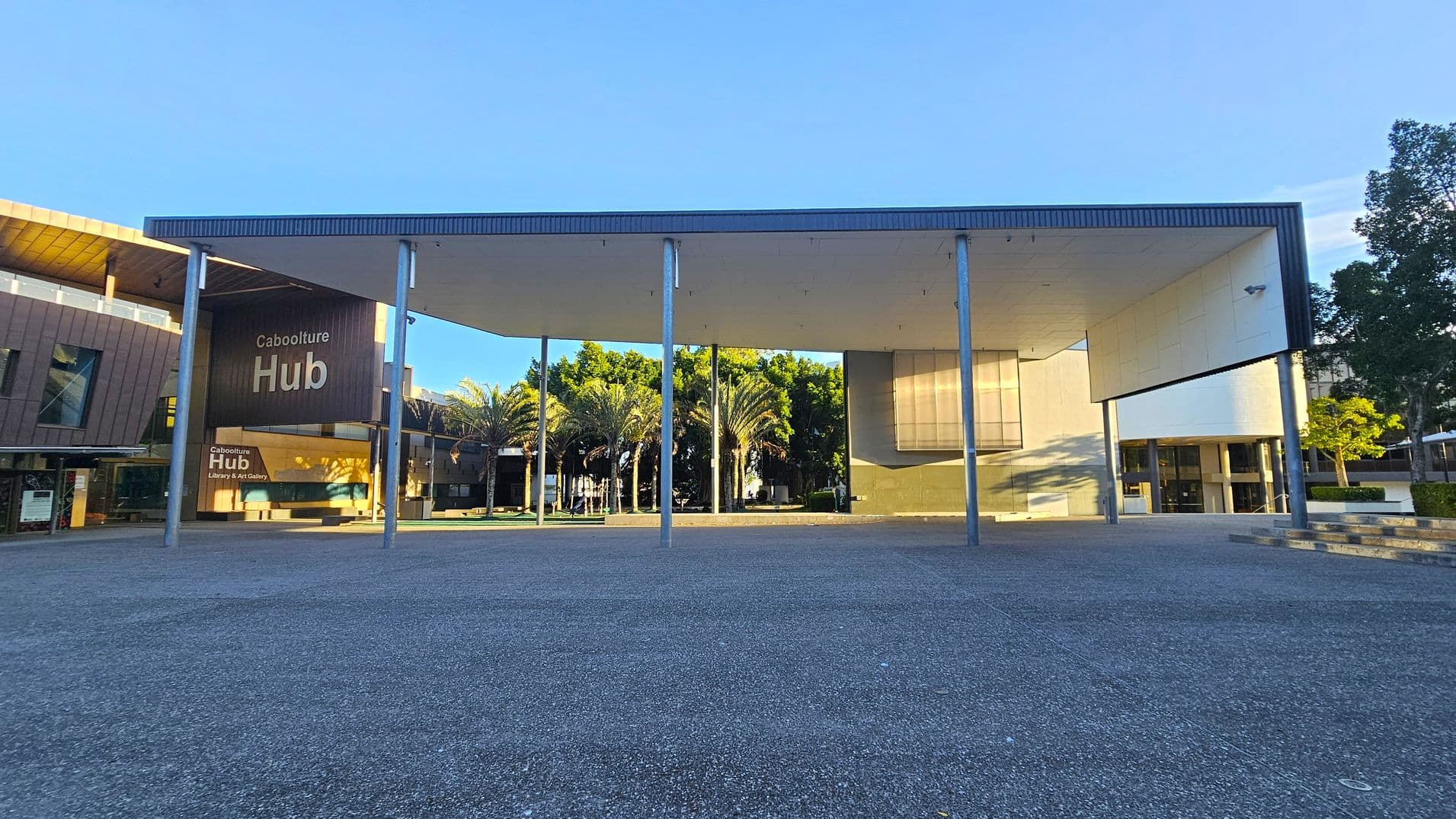 A modern building with a large, flat roof supported by tall metal columns. On the left, a sign reads "Caboolture Hub" with "Library & Art Gallery" below. The structure has a mix of glass and wood panelling. In the centre, palm trees are visible behind the columns. On the right, a white wall with a window and steps leading up to the entrance. The ground is paved with grey concrete, and trees are in the background.
