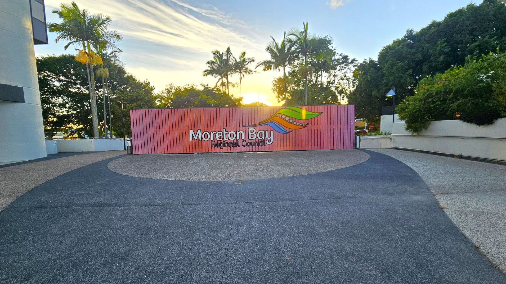 A large pink sign with "Moreton Bay Regional Council" and a colourful logo is in the centre. Tall palm trees and dense greenery are in the background, with the sun setting behind them. The foreground features a curved pathway with a mix of dark and light grey paving. On the left, part of a building is visible, while the right side has more greenery and a white wall.