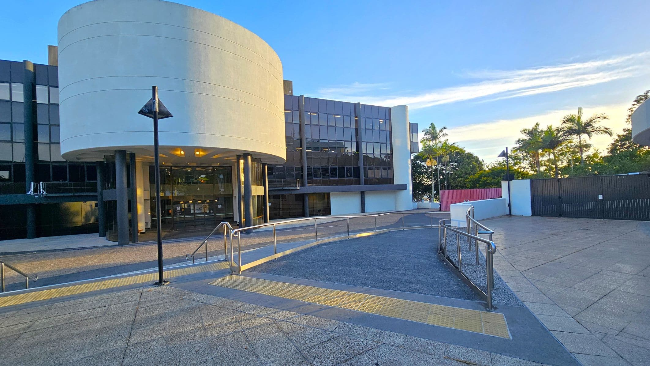 A modern building with a large, curved white facade on the left, featuring glass doors and windows. A ramp with metal railings leads up to the entrance. The ground is paved with textured tiles, and there are several black lamp posts. On the right, there are palm trees and a black fence. The background includes more trees and a clear sky.