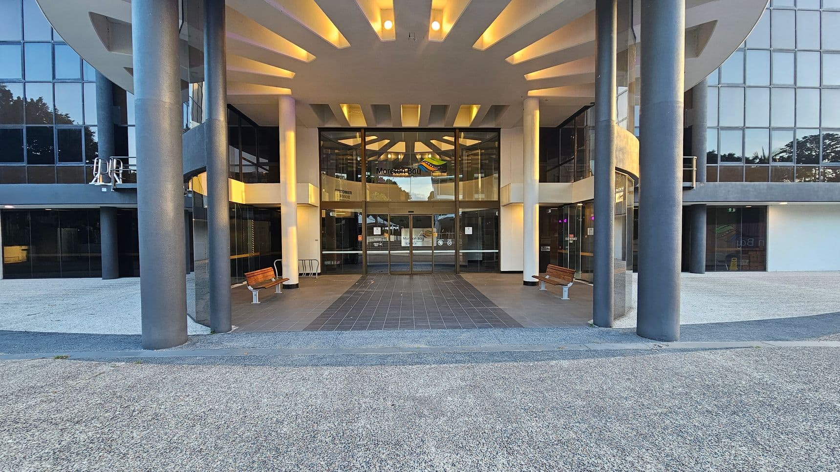 A modern building entrance with large glass doors in the centre, flanked by tall, dark grey columns. The ceiling features a geometric pattern with recessed lighting. Two wooden benches are positioned on either side of the entrance. The ground is covered with grey tiles leading to the doors, surrounded by a gravel area. The building facade is made of reflective glass panels, and there are trees visible in the reflection.