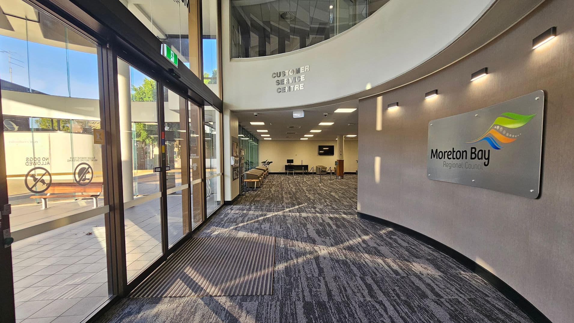 On the left, there are large glass doors with signs indicating "No Dogs Allowed" and a view of benches outside. The floor is tiled near the entrance and transitions to carpet inside. On the right, a curved wall displays a sign for "Moreton Bay Regional Council" with modern lighting above. In the centre, the space opens into a customer service area with chairs and a desk. The ceiling is fitted with recessed lighting, and a sign above reads "Customer Service Centre".