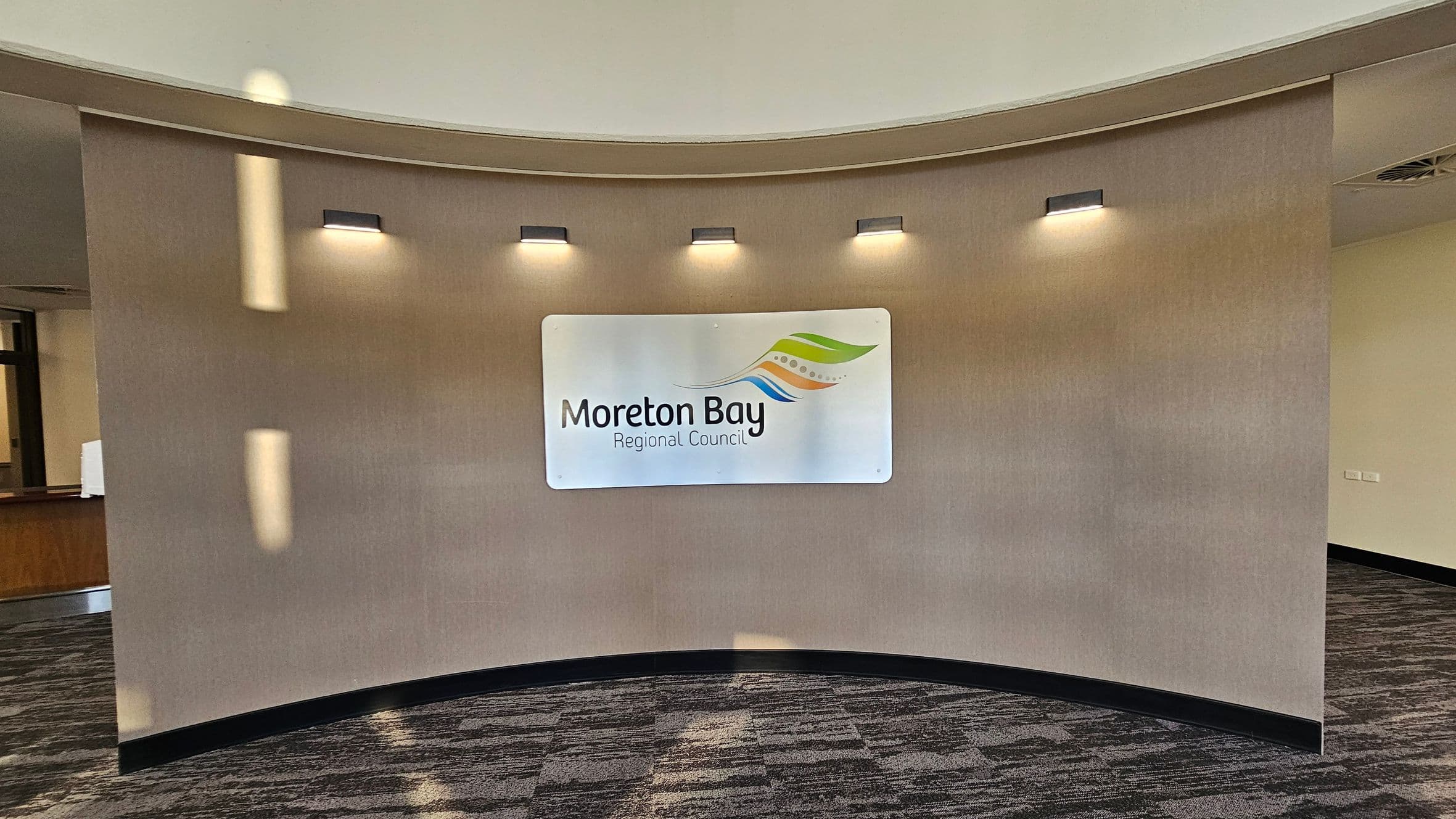 A curved wall with a beige finish features a sign in the centre that reads "Moreton Bay Regional Council" with a colourful logo above. Five small lights are evenly spaced above the sign, casting light downwards. The floor is carpeted with a pattern of grey and muted colours. To the left, a partial view of a wooden counter is visible, and to the right, the wall continues with a small section of a power outlet. The ceiling is light-coloured and curved.