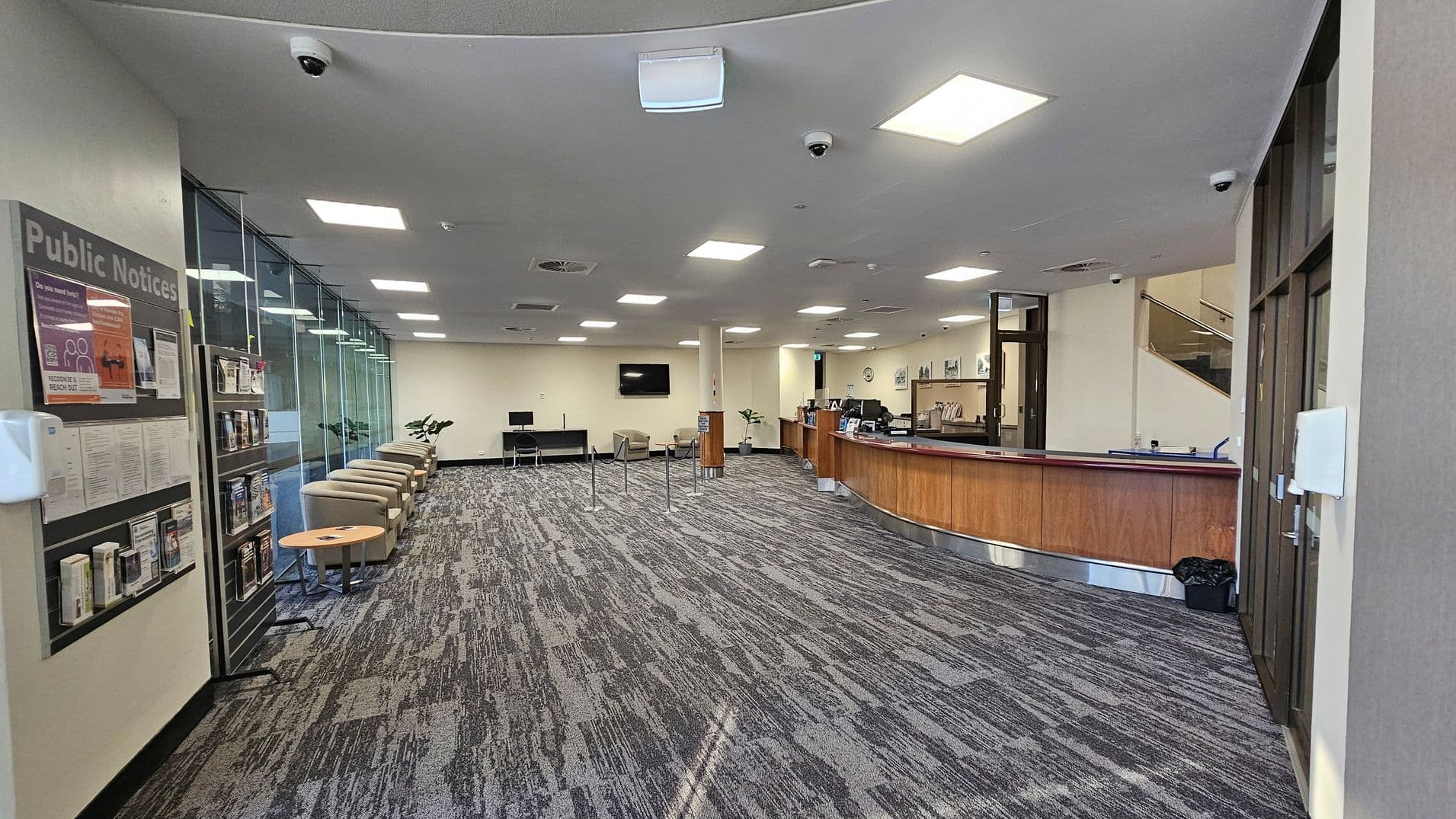 A spacious interior with a patterned carpet floor. On the left, a public notice board and a row of beige chairs with small tables are positioned along a glass wall. In the centre, a television is mounted on a cream wall above more seating. On the right, a curved wooden reception desk with a computer and various items. The ceiling is fitted with square lights and security cameras. In the background, a staircase leads to an upper level.