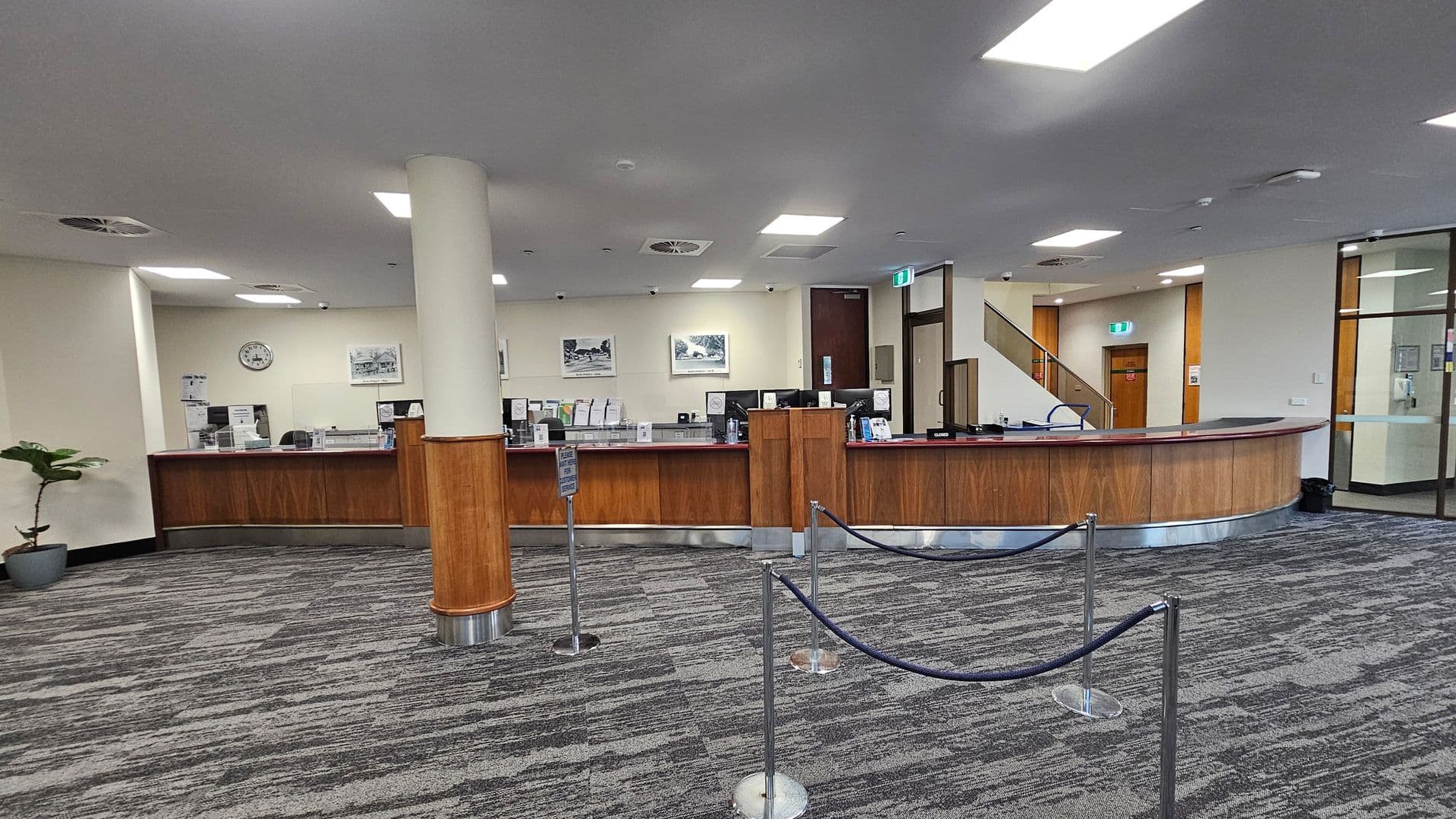 A spacious interior with a curved wooden reception desk in the centre, featuring multiple workstations and clear partitions. A light-coloured pillar stands in front of the desk. The floor is covered with grey patterned carpet. On the left, a potted plant is visible. The ceiling has recessed lighting and air vents. In the background, there are framed pictures on the wall, a clock, and a staircase leading upwards. A rope barrier is set up in front of the desk.