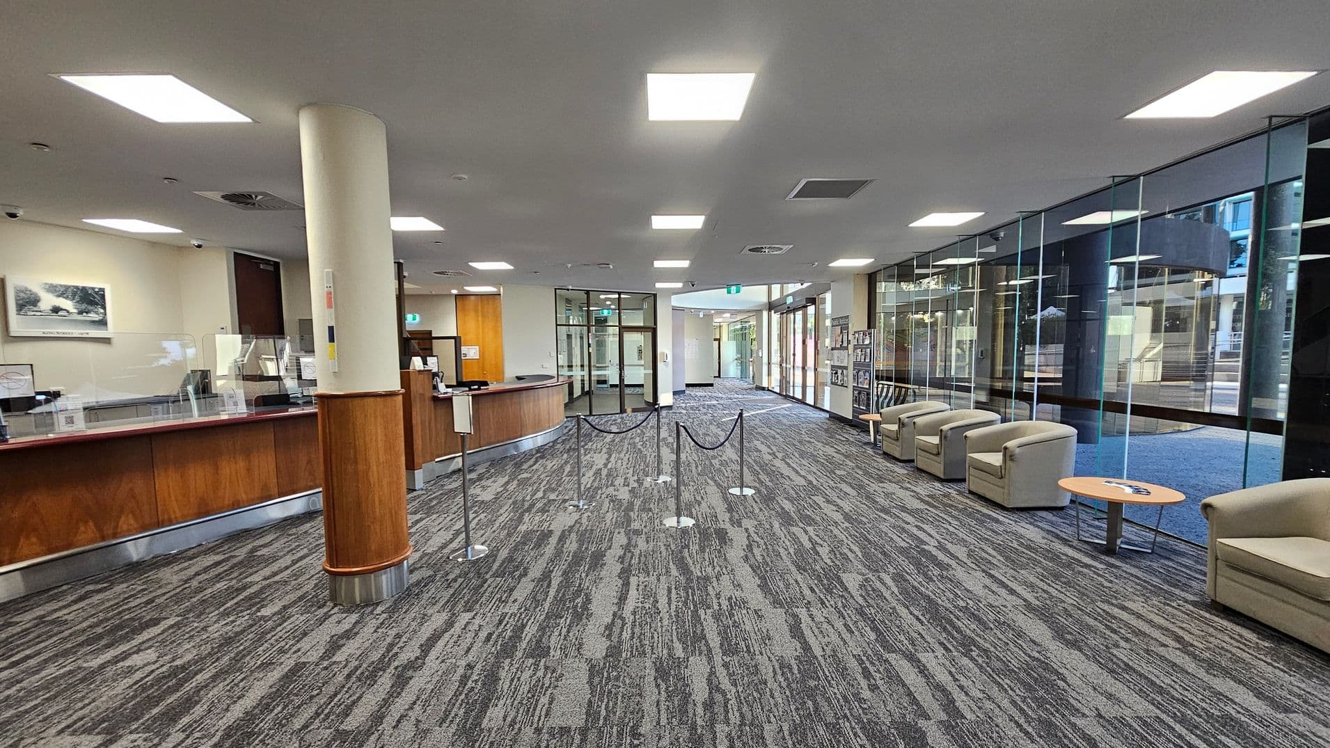 A spacious lobby area with a grey patterned carpet. On the left, there's a long wooden reception desk with glass partitions and a column. The right side features a row of beige armchairs with small round tables in front of a large glass wall. The ceiling has recessed lighting, and there are rope barriers in the centre of the room. The background shows glass doors leading to another area.
