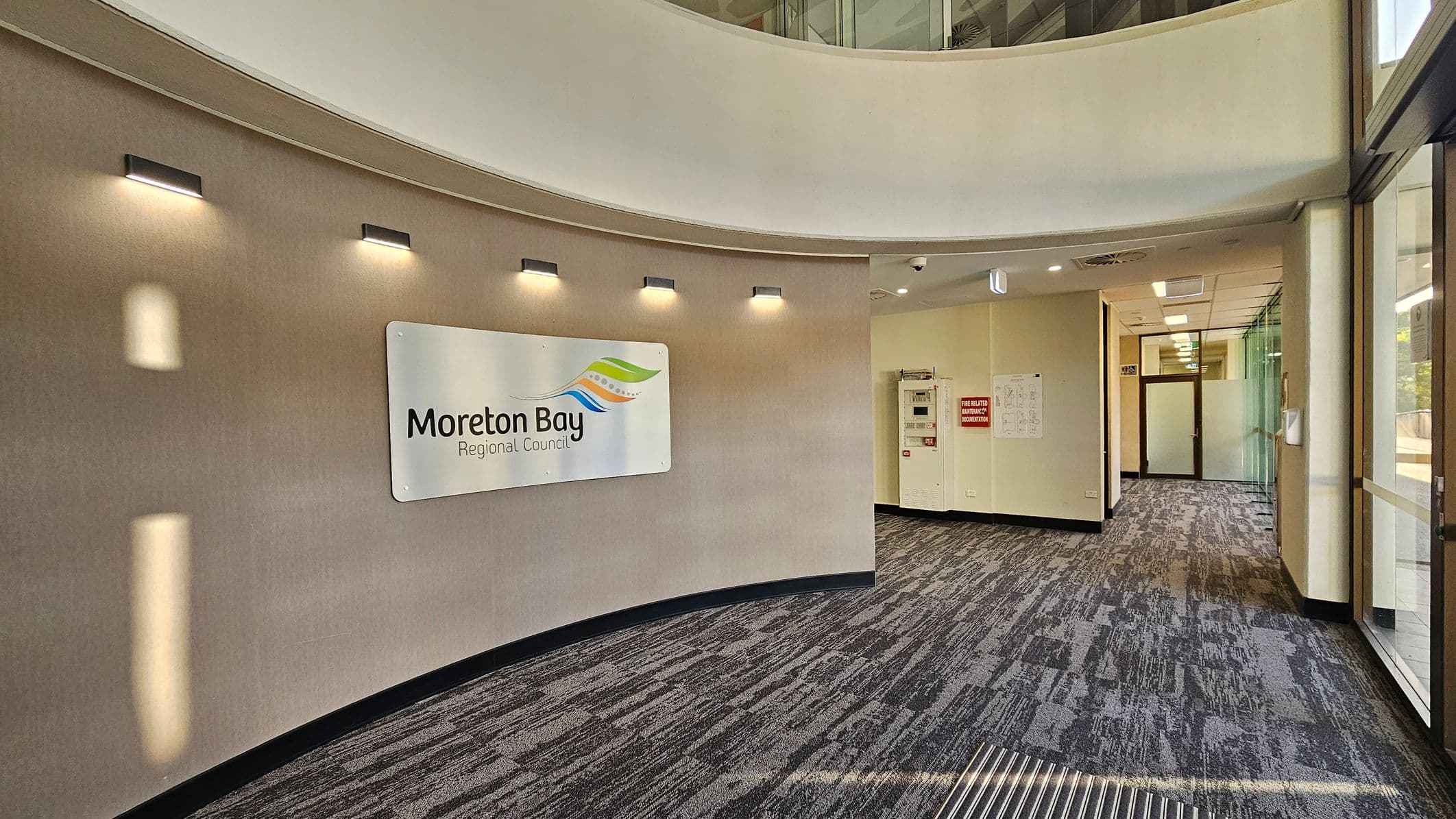 A modern interior space with a curved wall on the left featuring a sign for "Moreton Bay Regional Council" illuminated by several small lights. The floor is covered with a patterned carpet. On the right, there are large glass windows and doors. In the background, a hallway leads to a door with frosted glass panels. The ceiling has recessed lighting and ventilation panels.