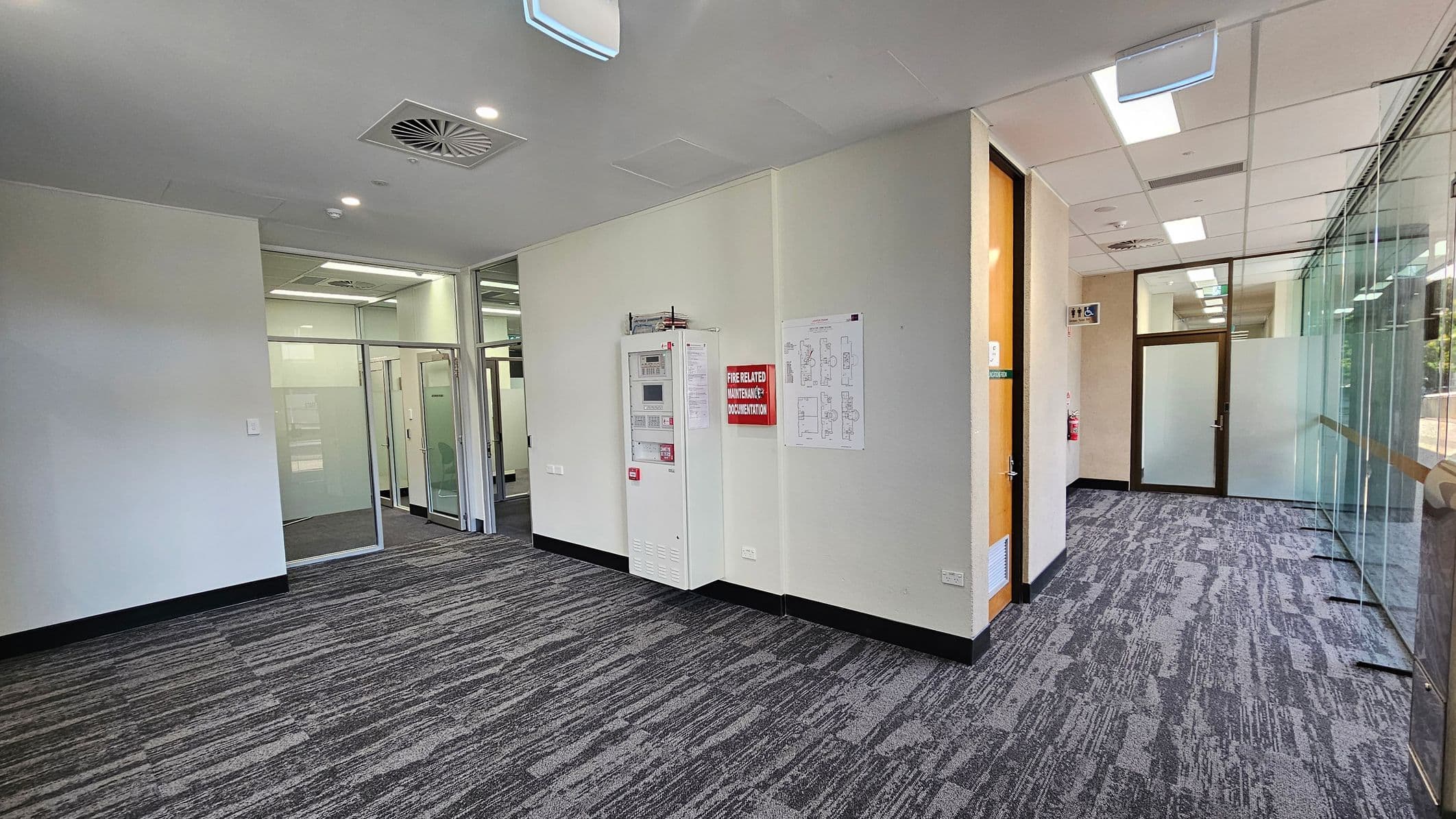 A modern office corridor with grey patterned carpet. On the left, a white wall features a fire control panel and a red sign. In the centre, glass doors lead to adjacent rooms. On the right, a glass wall with a view outside. The ceiling has recessed lighting and air vents. A wooden door is visible on the right side, and a fire extinguisher is mounted near the end of the corridor.
