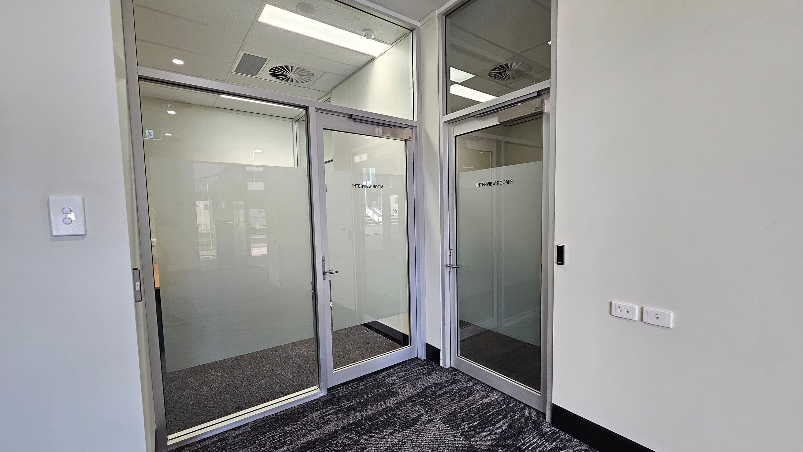 A hallway with two glass doors labelled "Interview Room 1" and "Interview Room 2" on the right. The doors have frosted lower panels and metal handles. The walls are light-coloured, and there are two light switches on the right wall. The floor is carpeted in a dark pattern. The ceiling features recessed lighting and air vents.