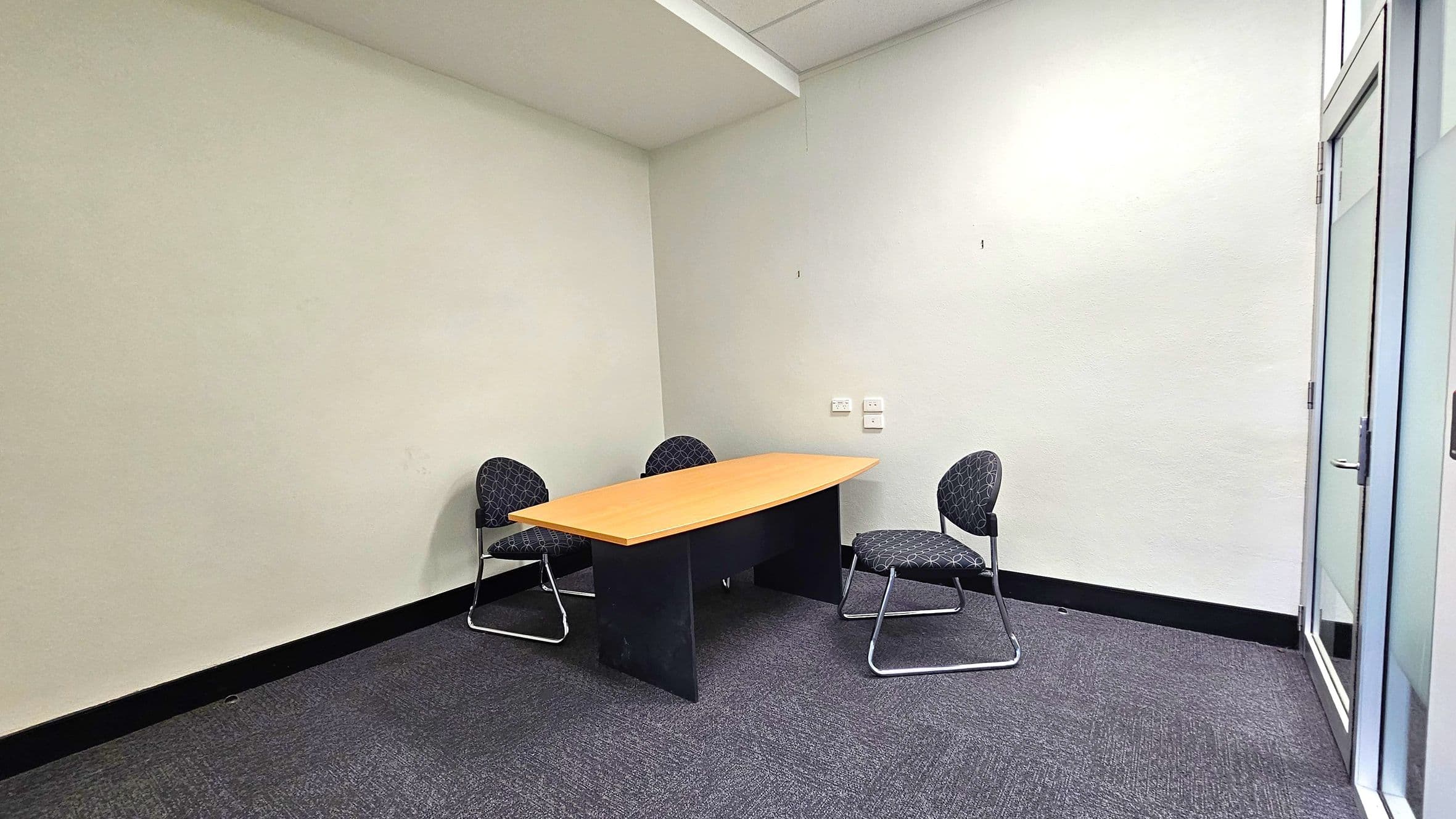 A small office room with a light grey carpeted floor and white walls. In the centre, there is a wooden table with a black base, accompanied by three black chairs with a circular pattern. The chairs have metal legs. On the right, there is a glass door with a metal handle. The ceiling is plain with a visible beam. The room has a simple and minimalistic design.
