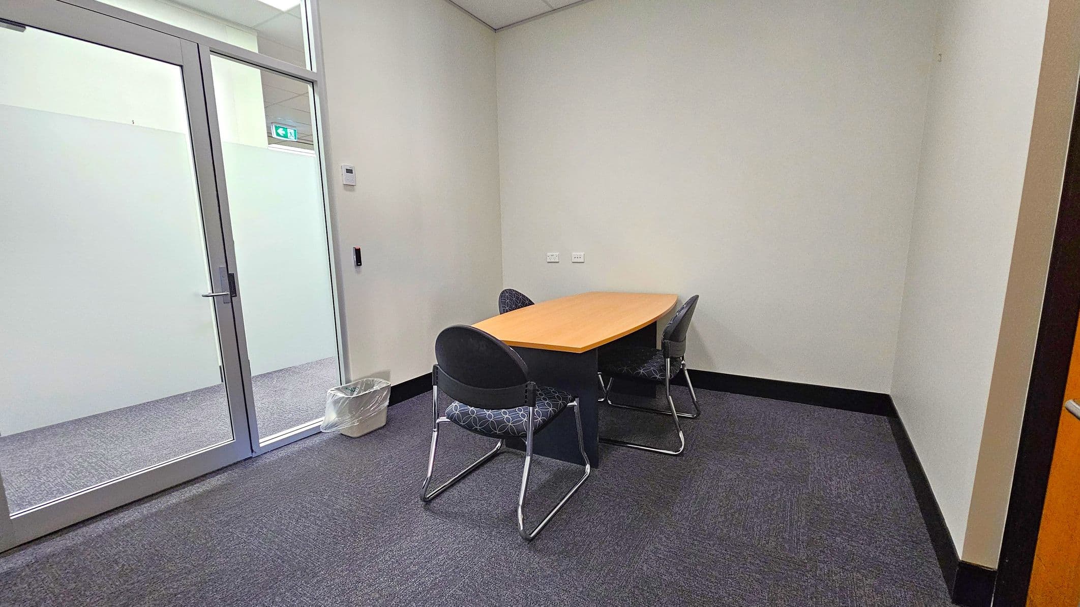 A small meeting room with a rectangular wooden table and three black chairs with patterned cushions. The floor is carpeted in grey. On the left, there is a glass door with a metal handle, and a small waste bin is placed nearby. The walls are plain and light-coloured, with a thermostat visible. The ceiling is white with recessed lighting.