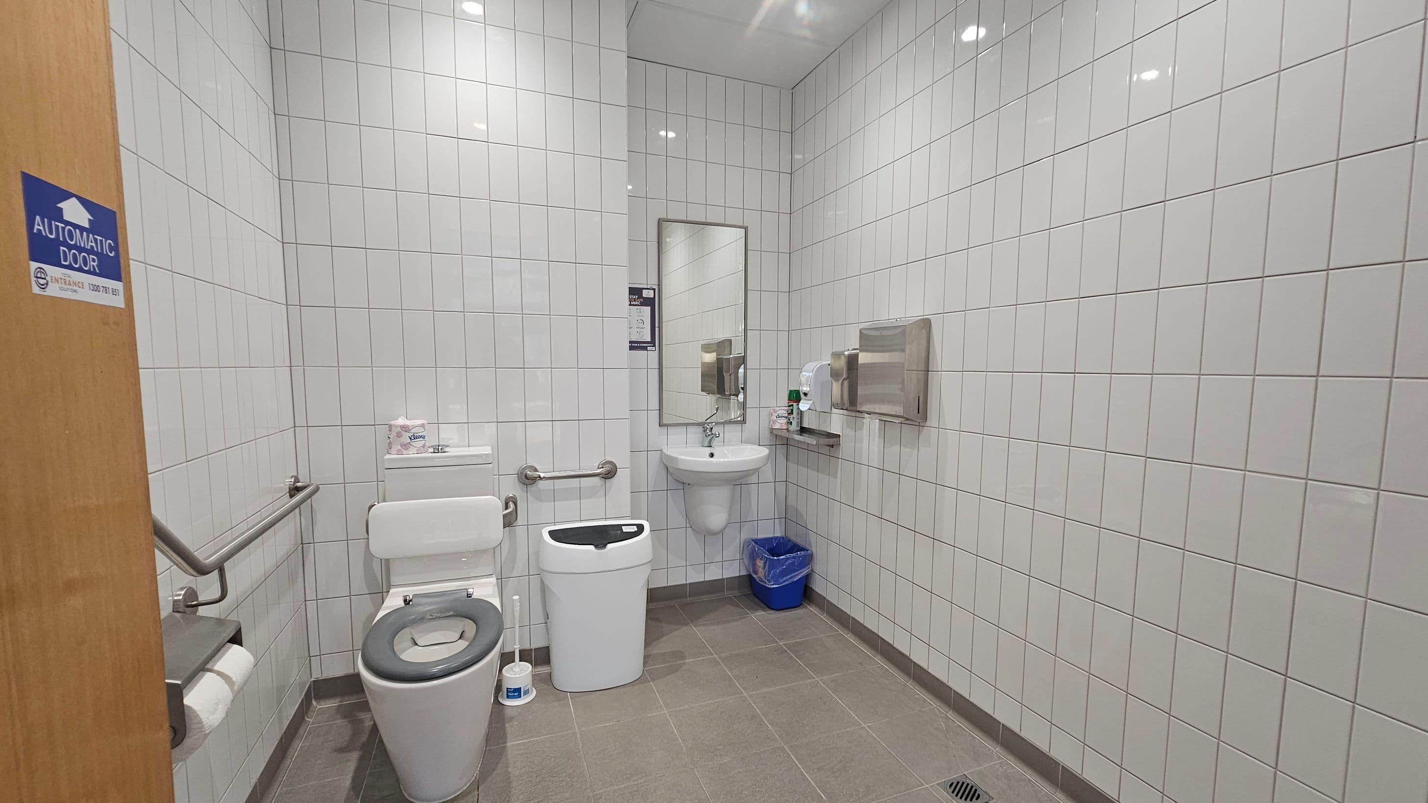 A public restroom with white tiled walls and a green tiled floor. On the left, a wooden door with a sign indicating an automatic door. A toilet with a black seat is next to a metal grab bar and a toilet roll holder. In the centre, a small sink with a mirror above it. To the right, a paper towel dispenser and a hand dryer are mounted on the wall. A blue waste bin is on the floor beneath the sink. The ceiling has recessed lighting.