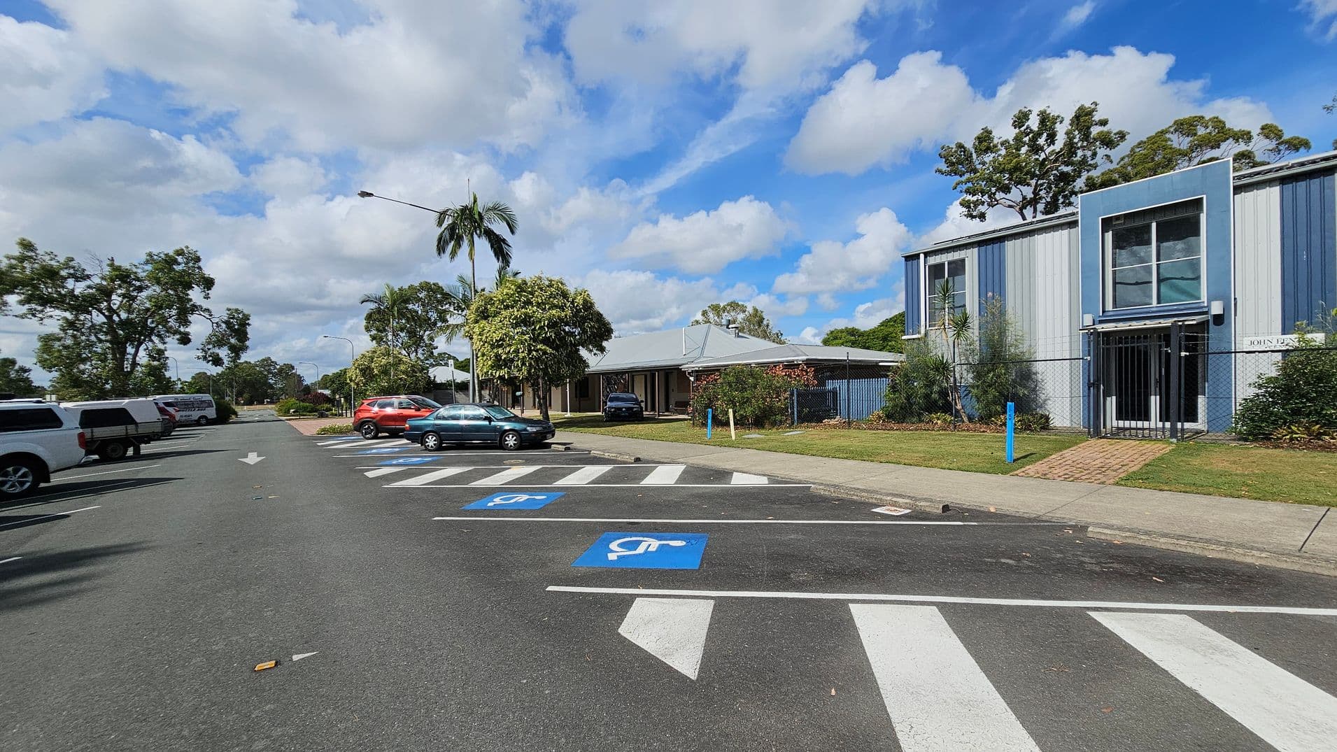 A street scene with a row of parked cars on the left, including a white pickup and a red SUV. On the right, there are designated disabled parking spaces marked in blue. A building with blue and grey panels is visible, featuring large windows and a gated entrance. Trees and shrubs are scattered throughout, with a palm tree near the centre. The road has a white arrow indicating direction, and a brick pathway leads to the building. The pavement is bordered by grass.