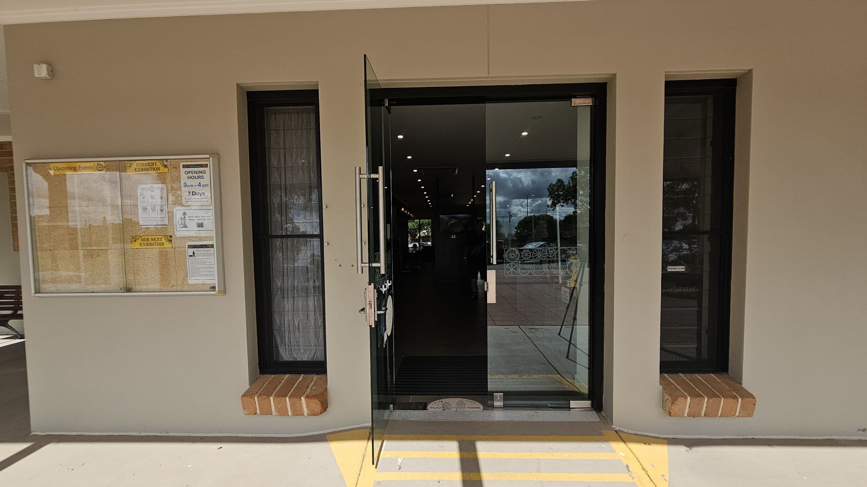 A glass door is centrally positioned, slightly open, with metal handles. On either side, there are narrow windows with black frames and brick sills. To the left, a notice board displays various papers and announcements, including opening hours. The floor is light-coloured with yellow lines leading to the entrance. Inside, ceiling lights are visible, and outside, trees and a paved area can be seen through the glass.
