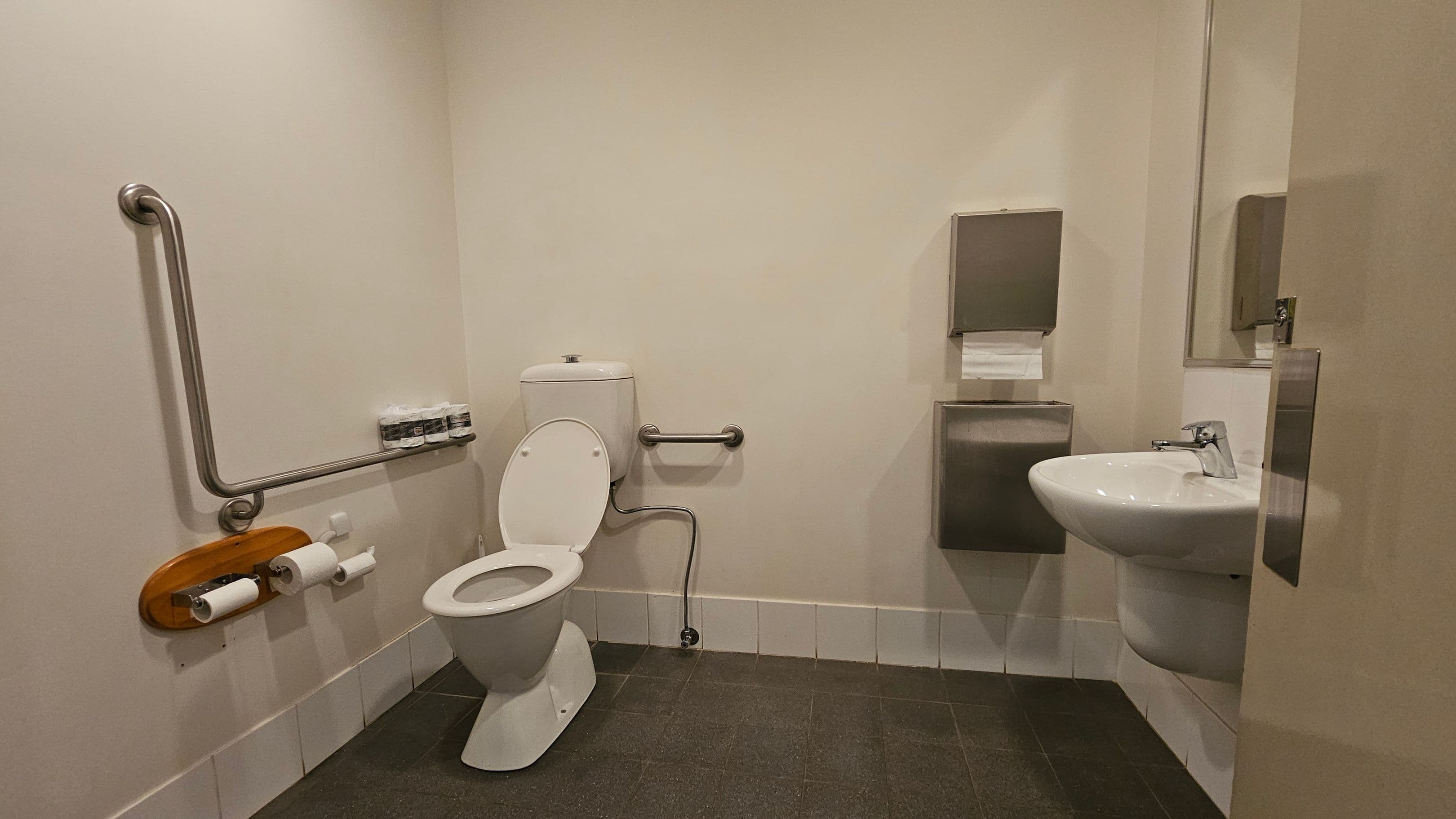 A bathroom with a white toilet on the left, featuring a metal grab bar and a wooden toilet paper holder with two rolls. Above the toilet, there are small bottles on a shelf. On the right, a white sink with a metal tap is mounted on the wall, accompanied by a metal paper towel dispenser. The floor is covered with dark tiles, and the walls are light-coloured. A partially open door is visible on the right side.