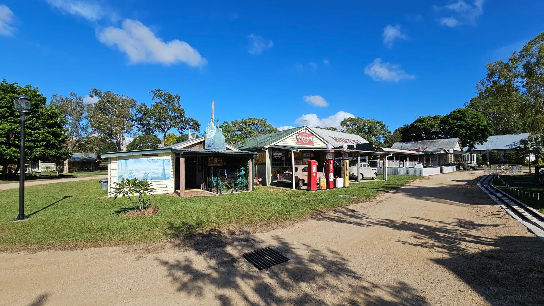 A rural scene with a dirt path running through the centre. On the left, a small building with a mural and a lamp post. In the middle, a larger building with vintage petrol pumps and a classic car under a sign. On the right, more buildings with corrugated roofs and a grassy area. Trees are scattered throughout the background, and the sky is clear with a few clouds.