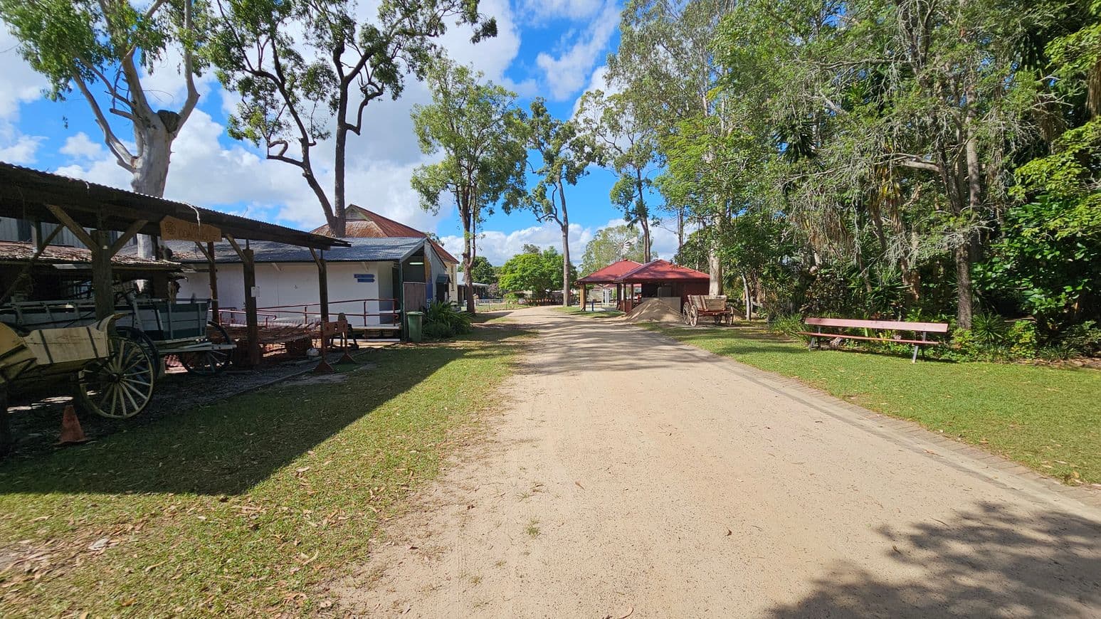 A dirt path runs through the centre, flanked by grass. On the left, there's a wooden structure with a roof, housing old wagons. To the right, a red-roofed shelter and a wooden bench are visible, surrounded by trees and greenery. Tall trees line the path, casting shadows on the ground.