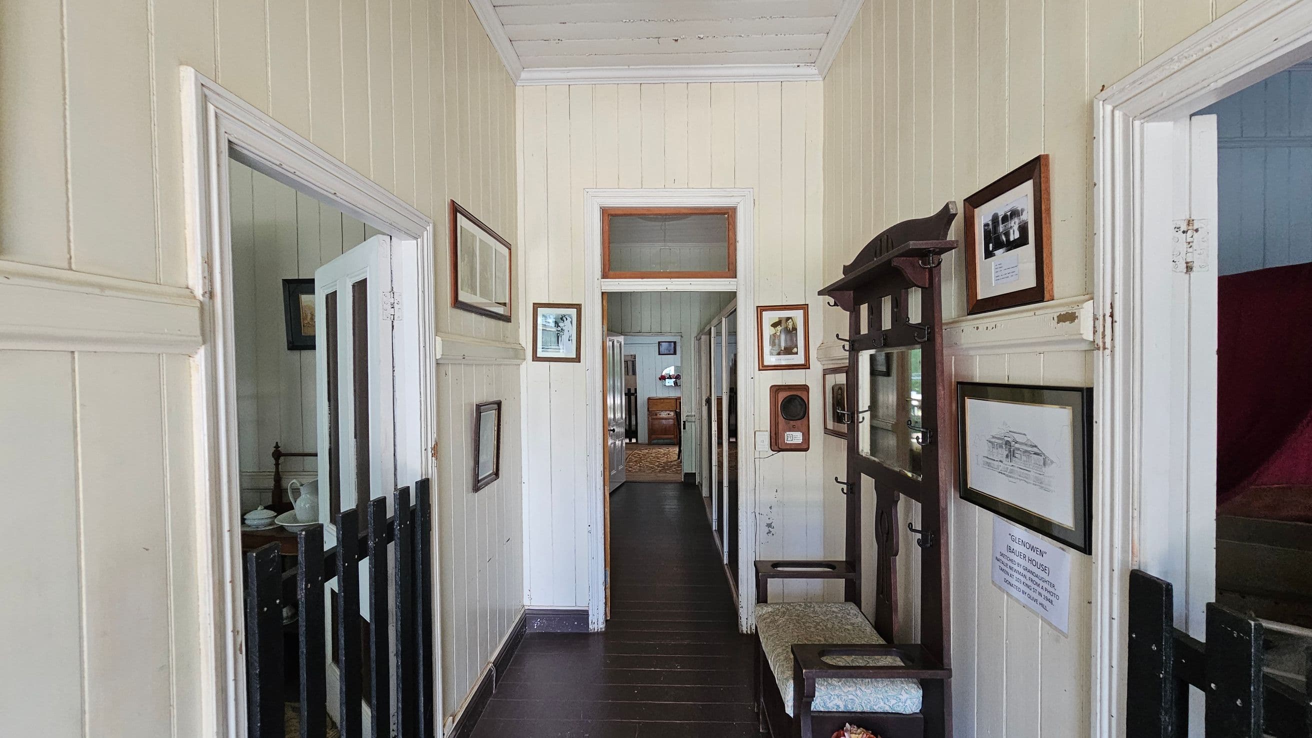 A narrow hallway with light-coloured wooden panelled walls and a dark wooden floor. On the left, an open door reveals a room with a bed and a small table. On the right, a wooden coat rack with a mirror and a cushioned bench is visible, along with framed pictures on the wall. At the end of the hallway, another open door leads to a room with a piece of furniture and a window. The ceiling is white and panelled.