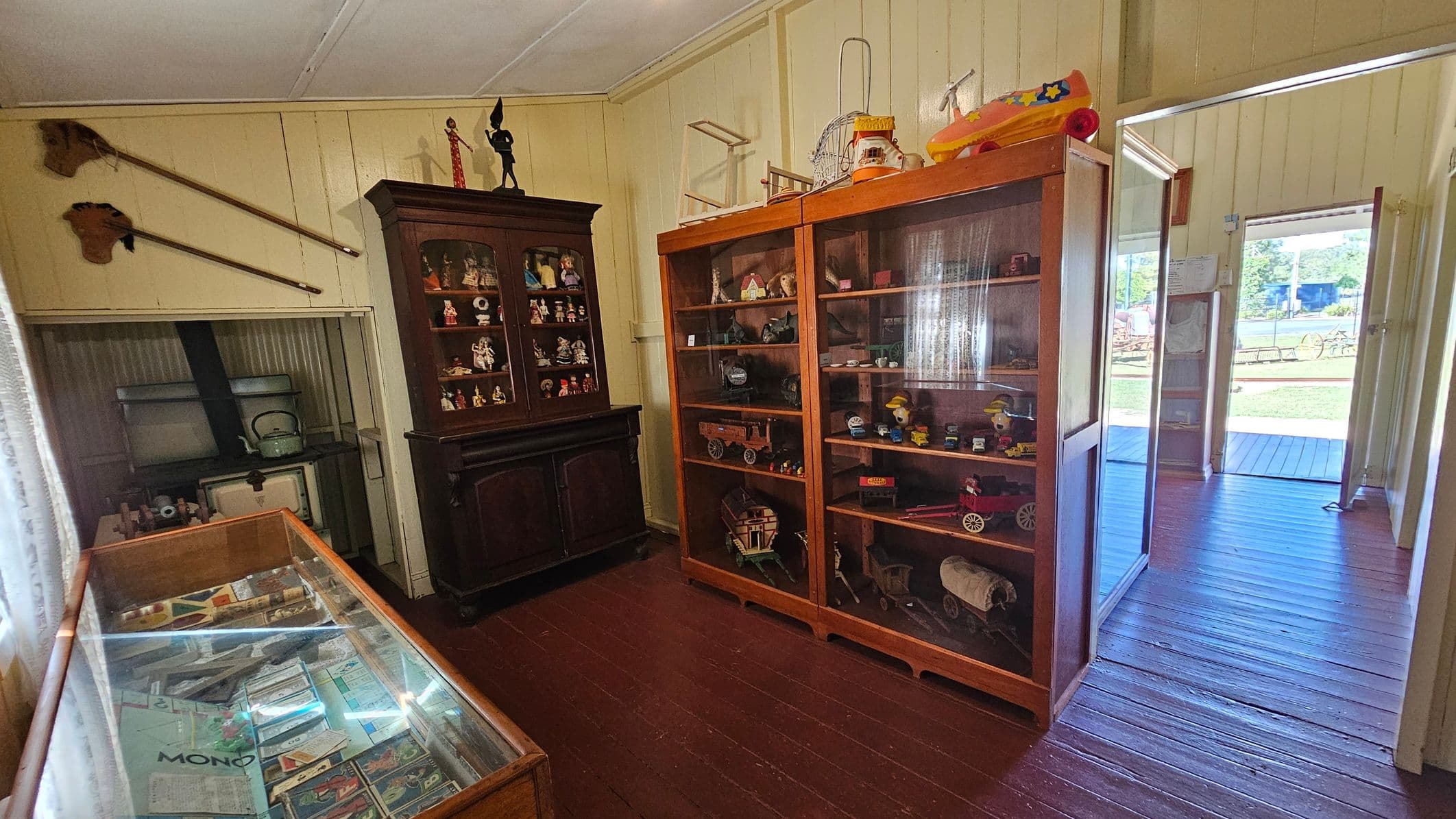 A room with wooden flooring and a white ceiling. On the left, a glass display case contains various documents and items. Above it, two long-handled tools are mounted on the wall. In the centre, a dark wooden cabinet displays small figurines. To the right, a large glass-fronted cabinet showcases vintage toys and models. The room opens to a porch with a view of a grassy area outside.