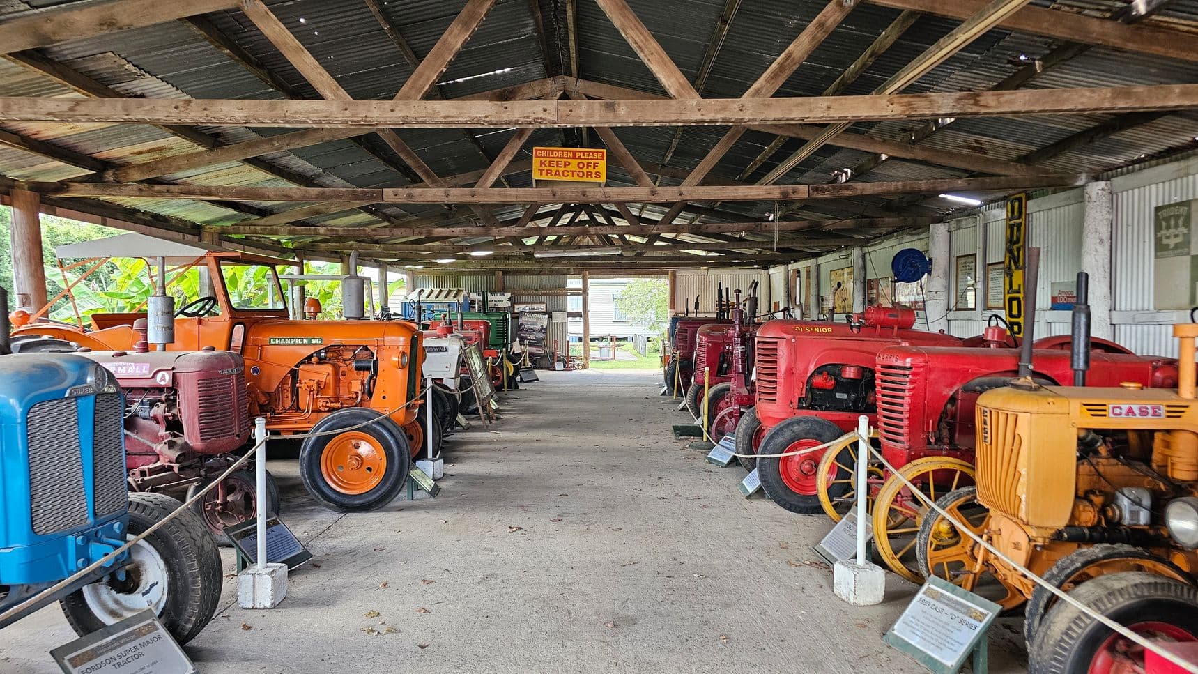 Inside a rustic shed with a corrugated metal roof supported by wooden beams, a collection of vintage tractors is displayed. On the left, there are blue and orange tractors, while on the right, red and yellow tractors are lined up. Each tractor is accompanied by an informational plaque. A sign hanging from the ceiling reads, "Children Please Keep Off Tractors." The floor is concrete, and the walls are lined with various signs and memorabilia. In the background, greenery is visible through the open end of the shed.