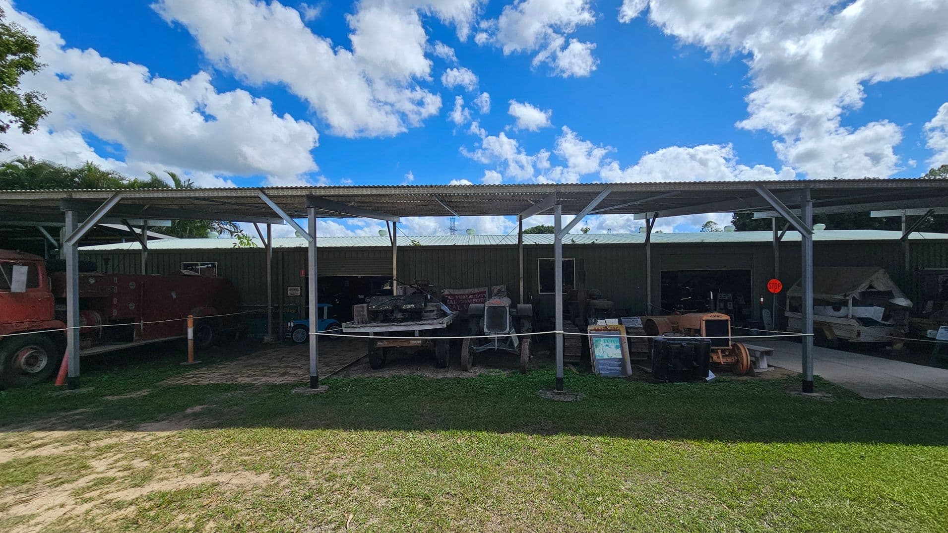 A covered outdoor area with a corrugated metal roof supported by metal beams. Underneath, various machinery and equipment are displayed, including a tractor and other vintage vehicles. A rope barrier separates the display from the grassy foreground. The background features a metal building with open sections revealing more equipment inside. A "STOP" sign is visible on the right. The ground is a mix of grass and dirt.